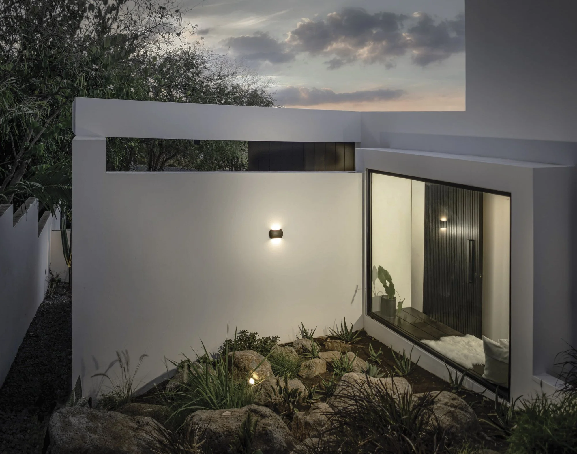 Une vue d'une façade de maison moderne blanche avec une grande fenêtre au rez-de-chaussée, un éclairage extérieur, un jardin avec des rochers et des plantes, et un ciel avec des nuages roses au coucher du soleil.