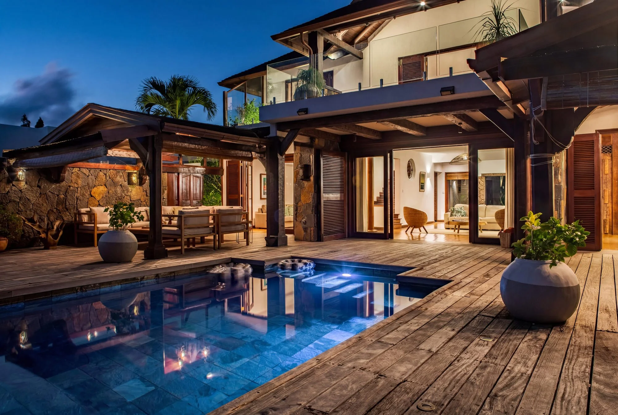 Maison moderne avec terrasse et piscine la nuit, intérieur éclairé visible à travers de grandes fenêtres, meubles en bois, plantes, décor chaleureux et ambiance détendue.