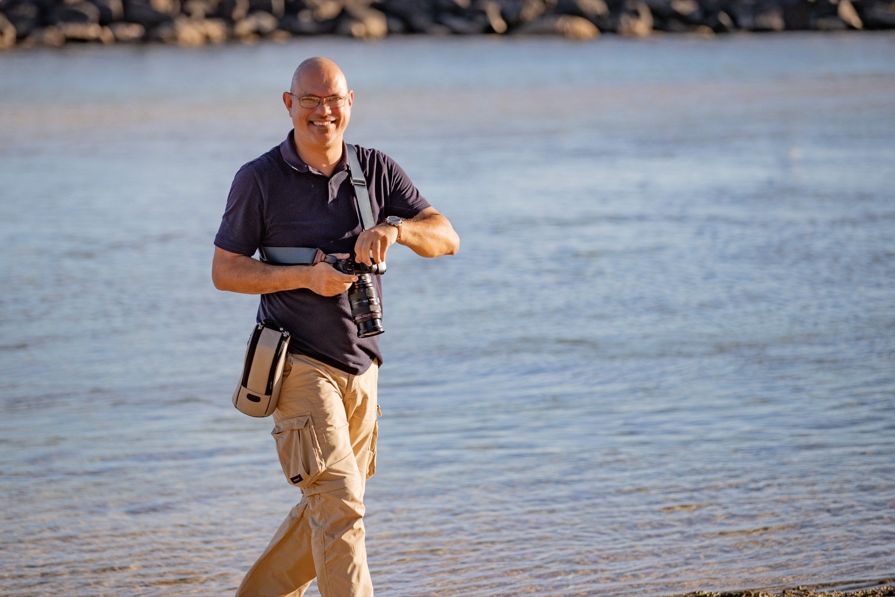 Homme souriant portant des lunettes, une caméra autour du cou, marchant sur la plage