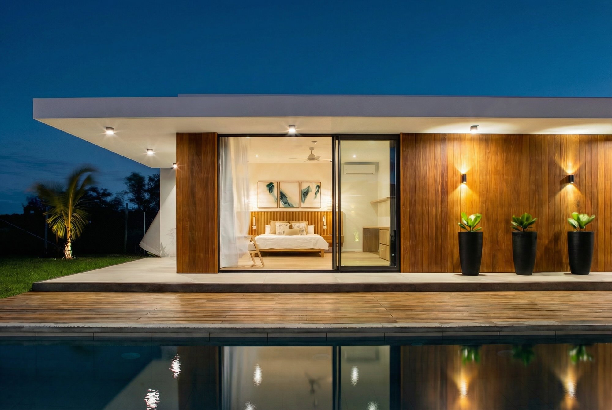 Maison moderne avec grande baie vitrée, éclairée la nuit, avec piscine reflétant la maison.