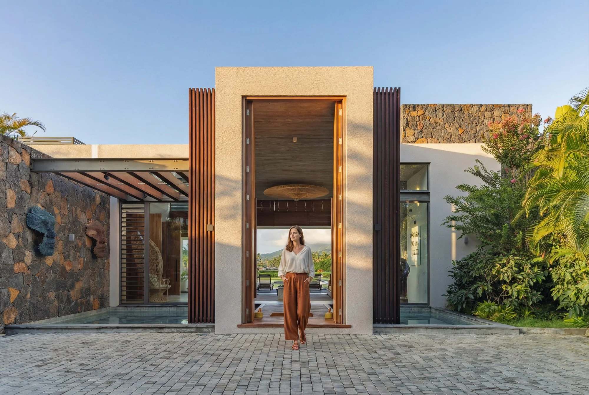 Une femme se tient devant une entrée moderne d'une maison, entourée de végétation luxuriante et d'un ciel clair, avec un design architectural contemporain.