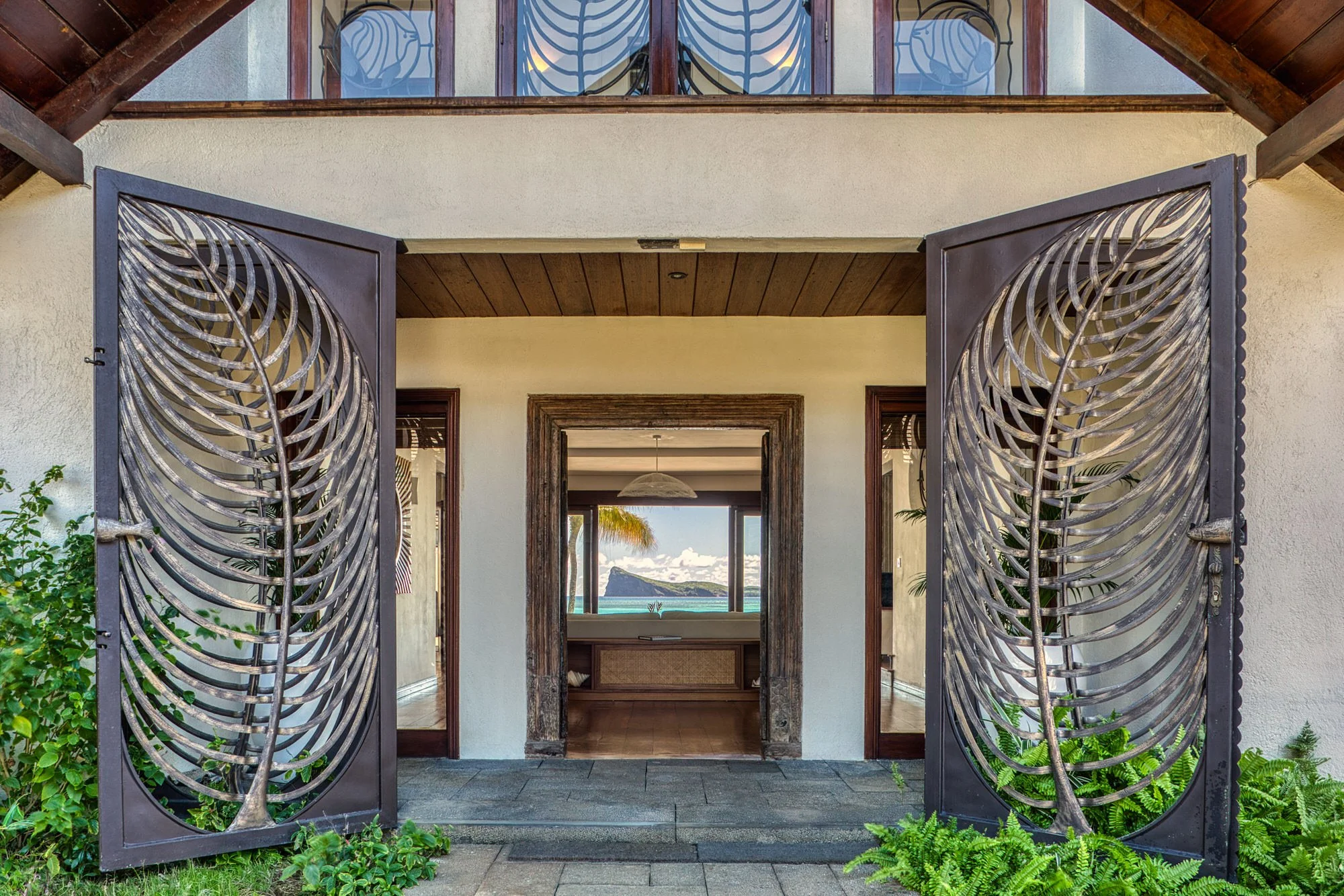 Portail en métal avec un motif de spirale en bois, ouvrant sur une maison avec vue sur l'océan, intérieur en bois, murs blancs, porte en bois sculpté, vue depuis l'entrée vers une pièce avec vue sur la mer.