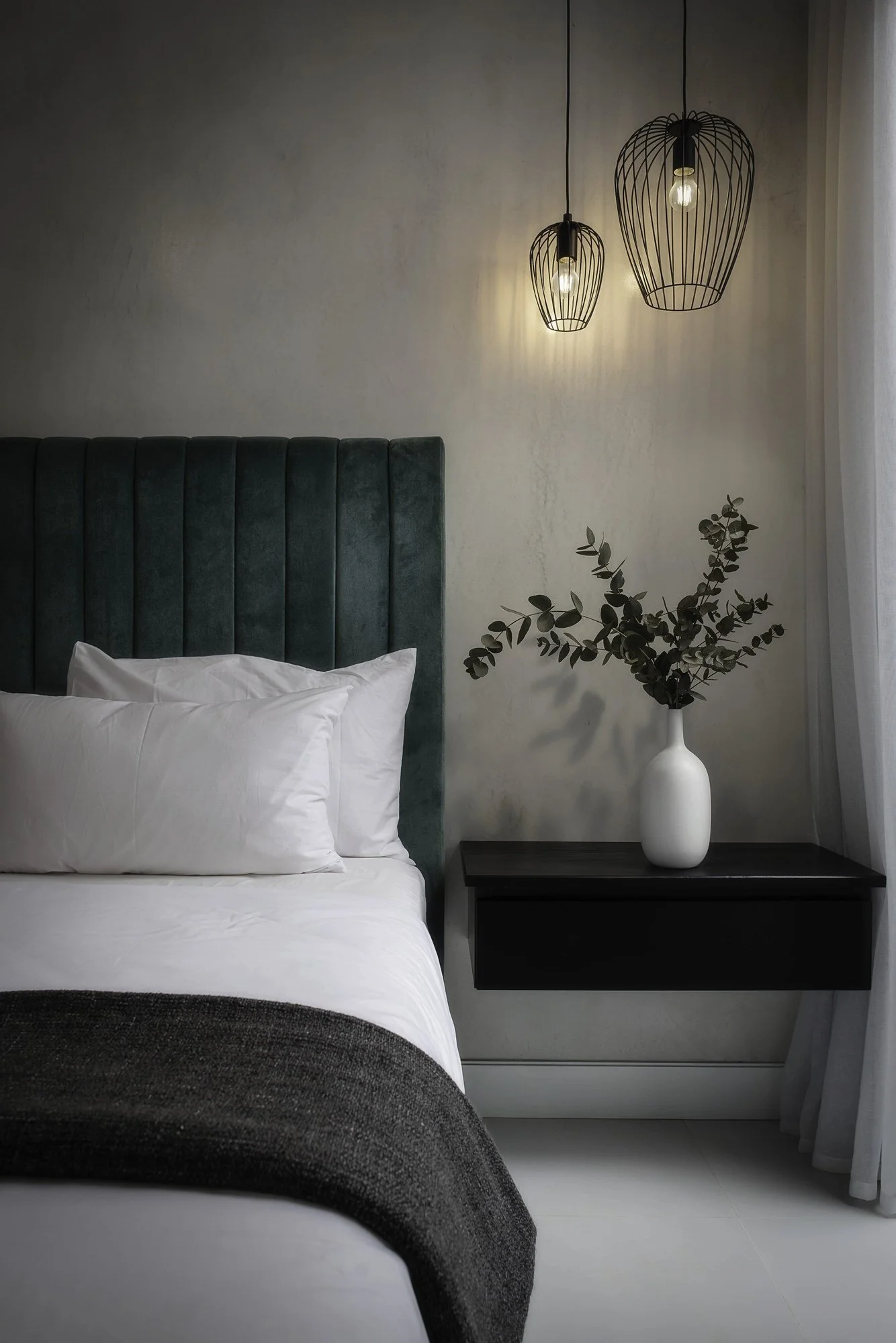 Un lit avec une tête de lit en velours vert, à côté d'une table de chevet noire avec une vase blanche contenant des feuilles de eucalyptus, et deux suspensions en fil de fer avec des ampoules, dans une chambre lumineuse.