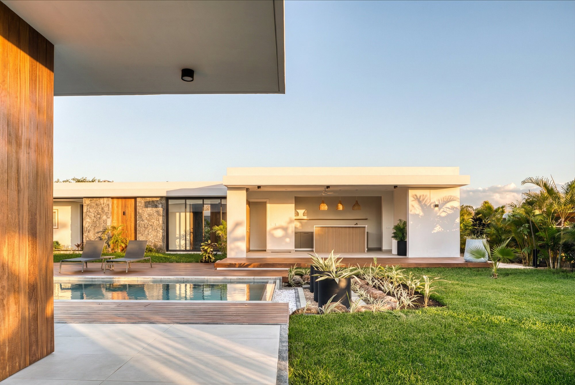 Vue d'une maison moderne avec une piscine, un patio en bois, un espace de vie extérieur, chaises longues, et un jardin verdoyant, sous un ciel clair.