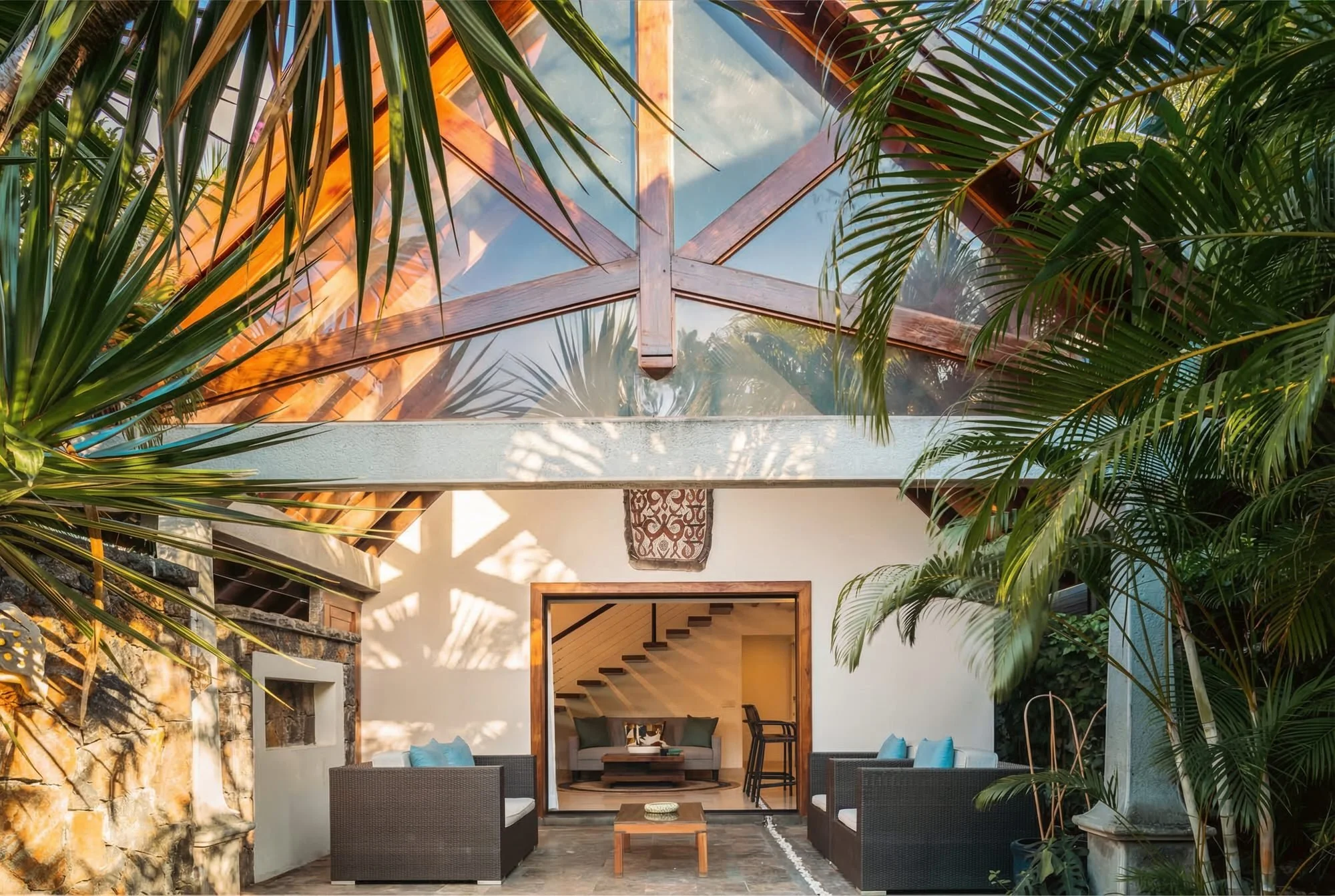 Salon de maison avec mobilier en bois et canapés, ouvert sur un espace extérieur avec plantes tropicales et toiture en verre