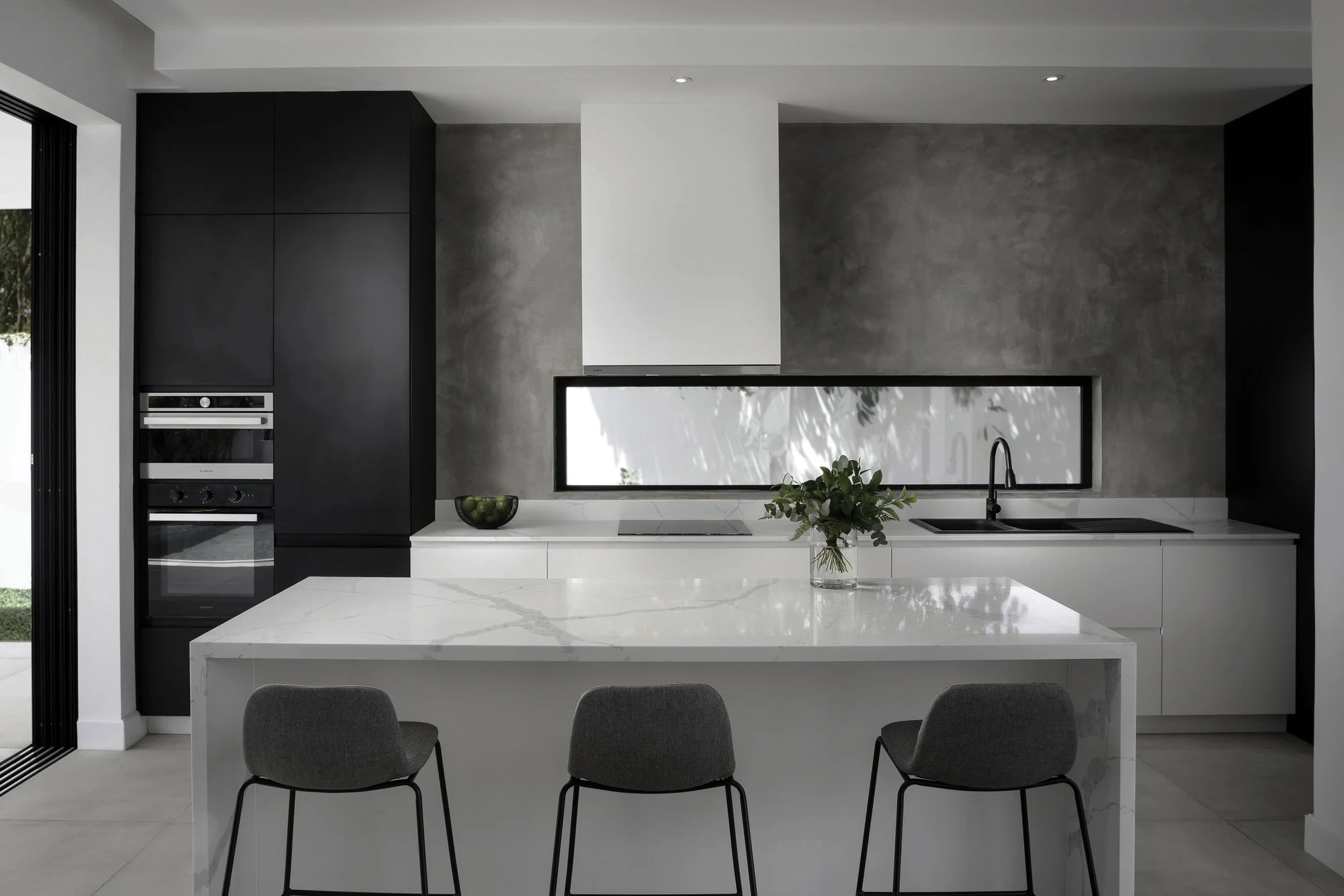 Cuisine moderne avec îlot en marbre blanc, murs gris et noirs, réfrigérateur encastré, évier noir, vase avec plantes, fenêtre large laissant entrer la lumière