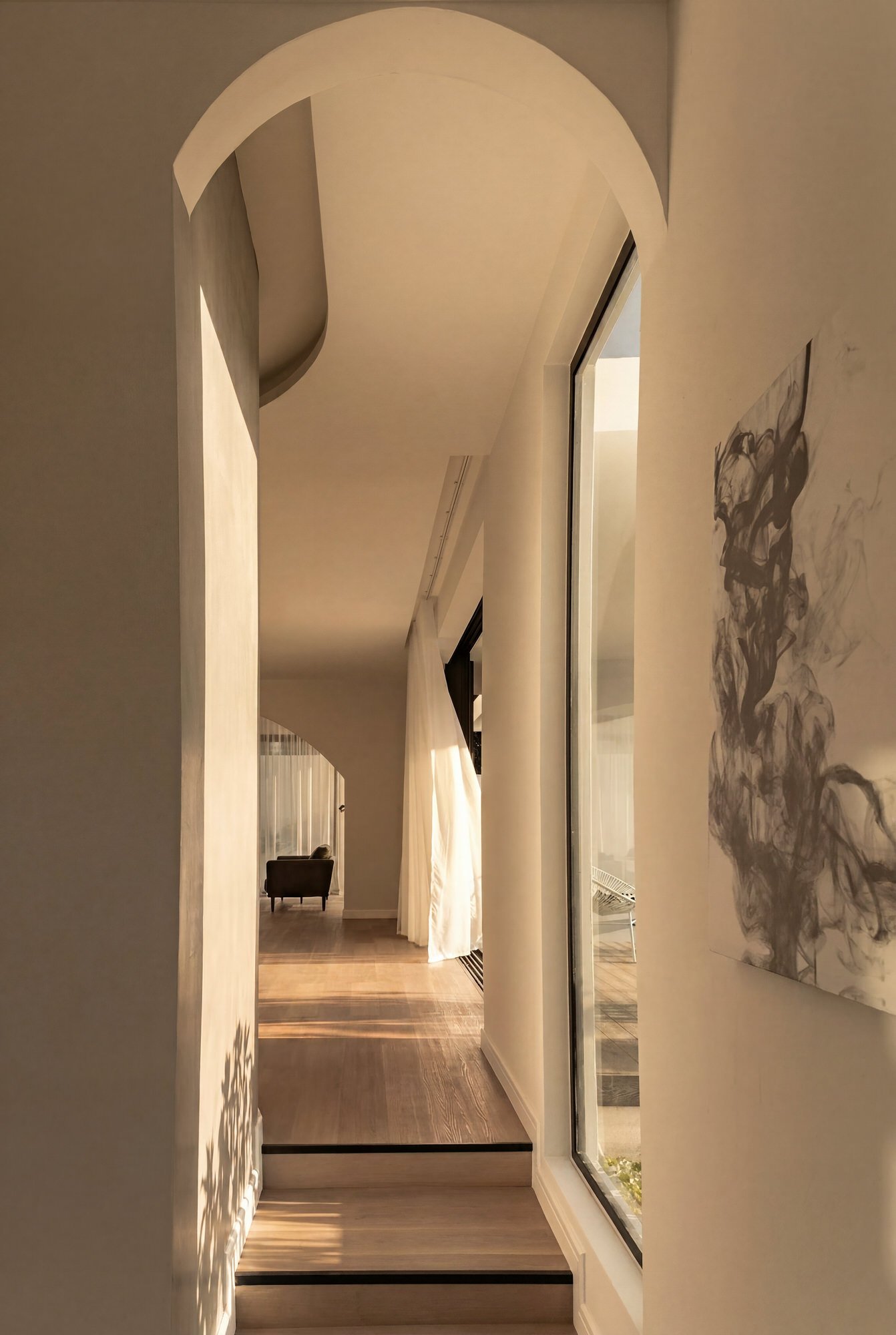 Vue intérieure d'un espace moderne avec un couloir, un mur blanc avec une peinture abstraite, des rideaux blancs, et un fauteuil noir à l'arrière, avec une architecture élégante et un éclairage chaud.