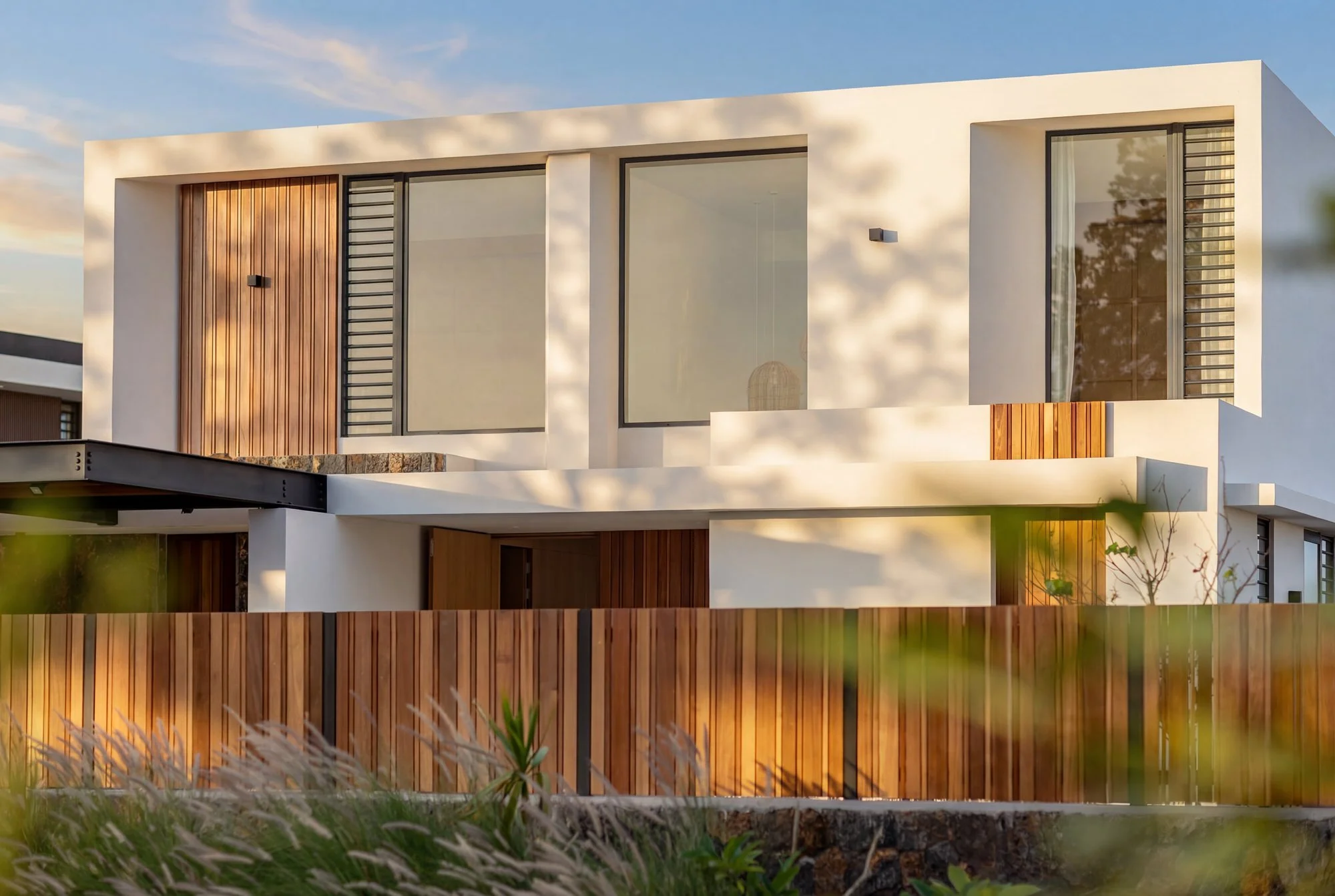 Maison moderne à deux étages avec grandes fenêtres, murs blancs, détails en bois et un jardin avec clôture en bois.