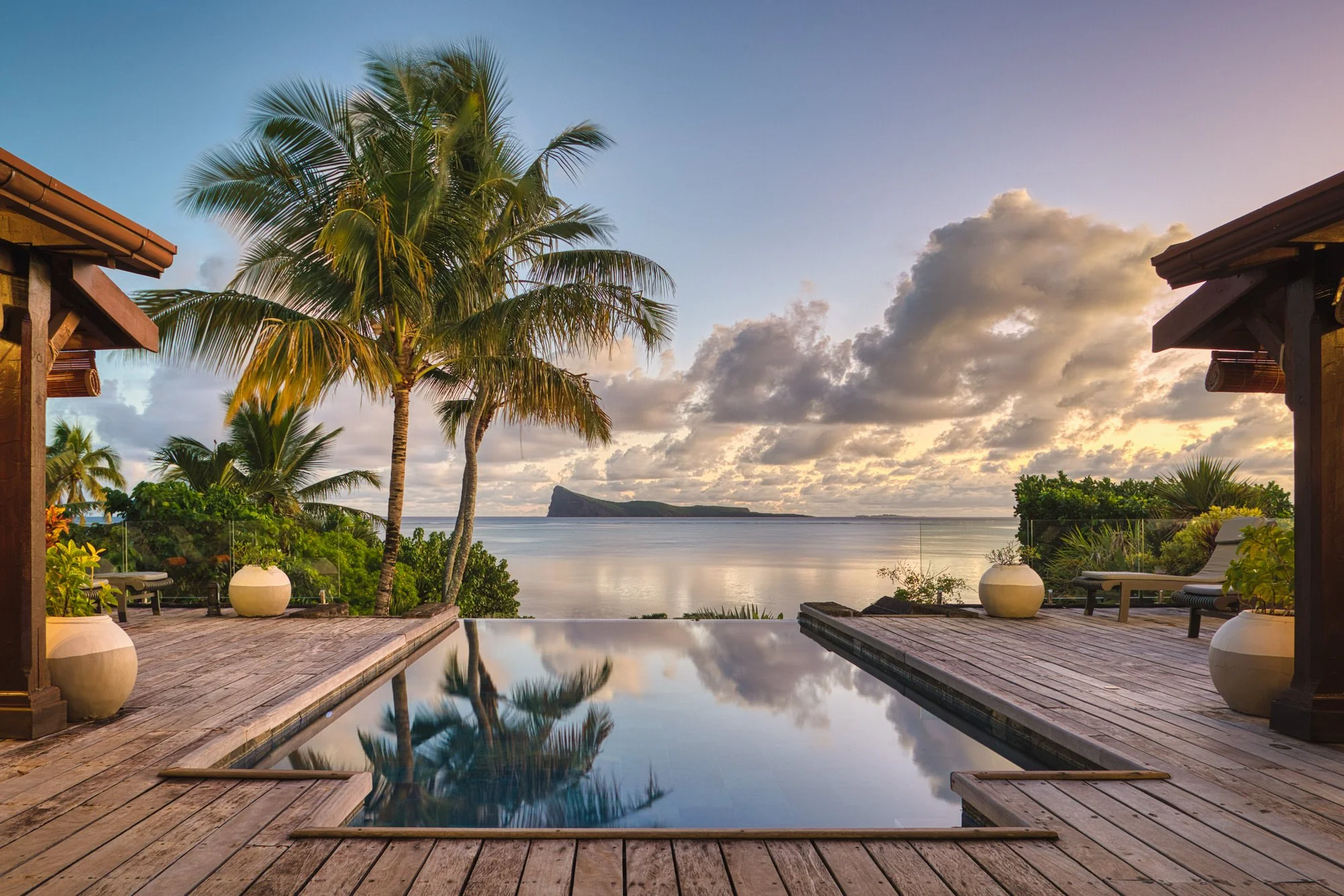 Une terrasse en bois avec un miroir d'eau reflétant un ciel de coucher de soleil, entourée de palmiers et de plantes tropicales, donnant une vue sur la mer avec une montagne à l'horizon.