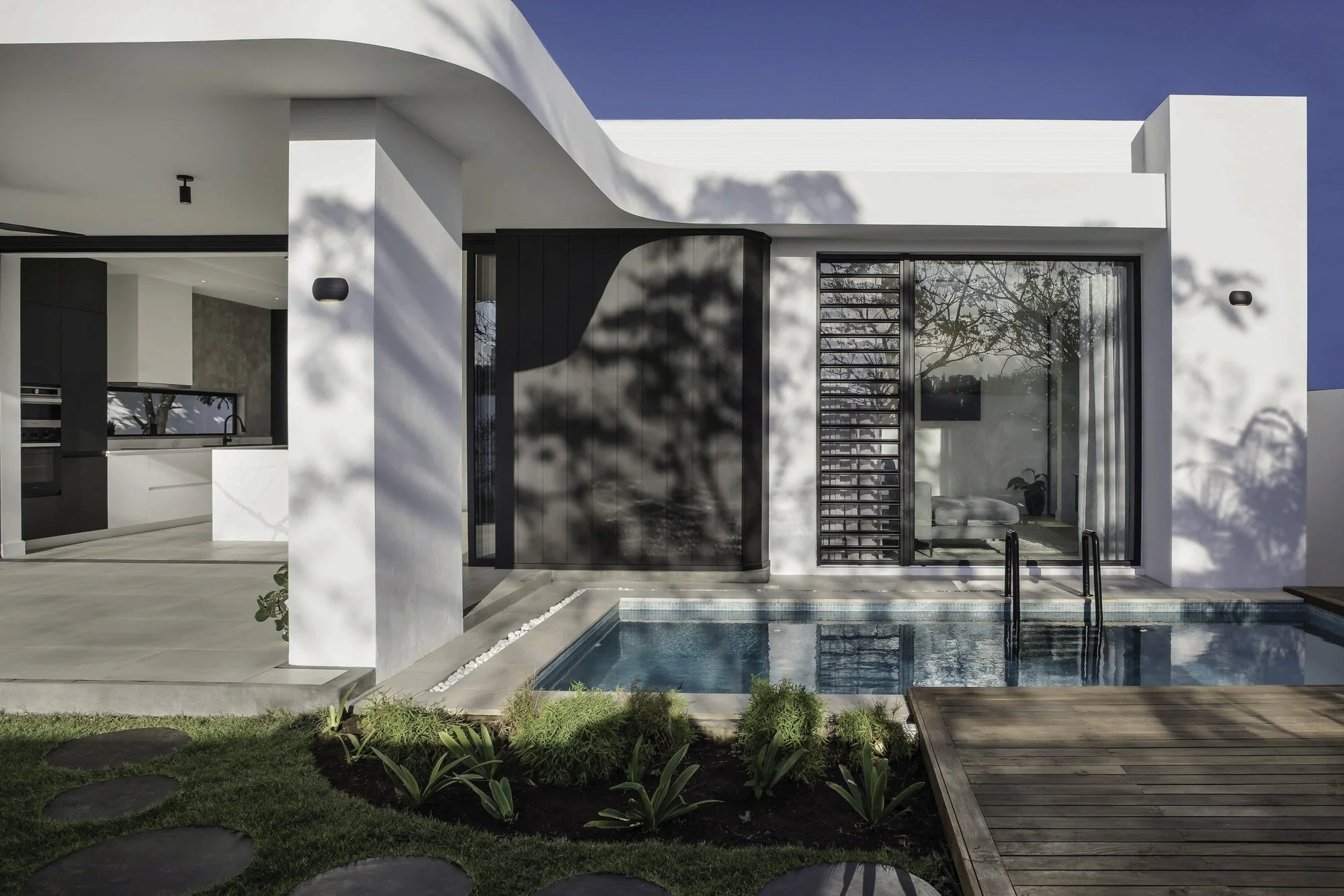 Façade moderne d'une maison avec piscine, grande fenêtre vitrée, espace extérieur avec dalles en bois et jardin avec plantes et galets