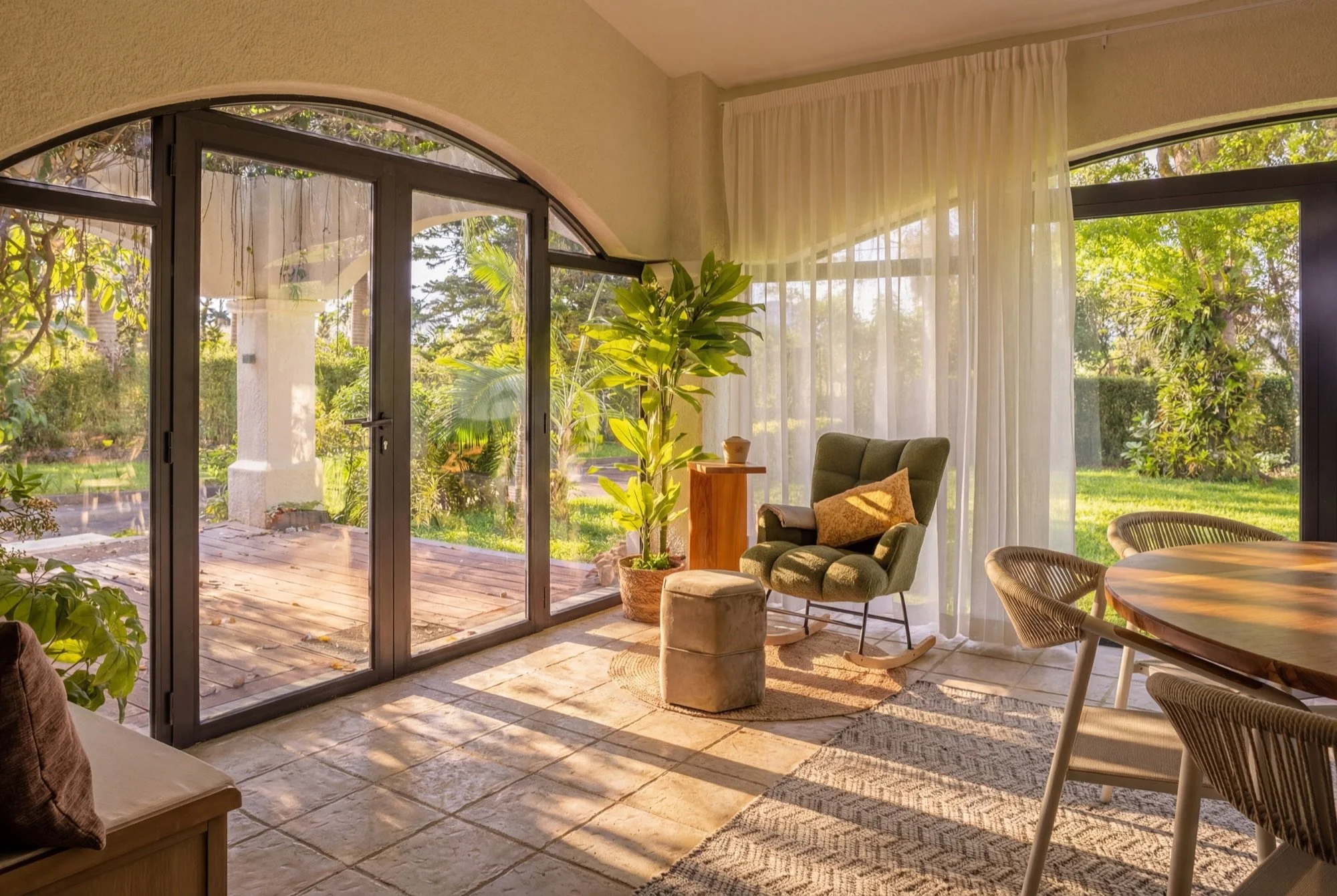 Salon avec grande baie vitrée, fauteuil vert, plante verte, table ronde en bois, rideaux blancs, vue de l'extérieur avec jardin et arbres.