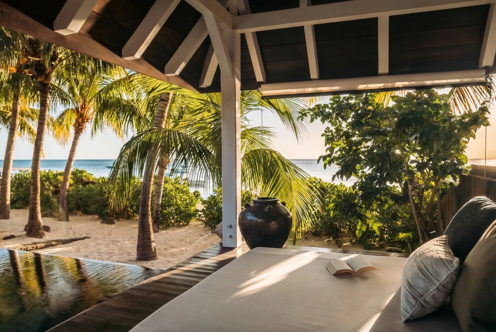 Une vue depuis une terrasse ou un patio avec un canapé et un livre ouvert, regardant des palmiers et la plage avec la mer au loin, sous un ciel en fin d'après-midi ou en début de soirée.