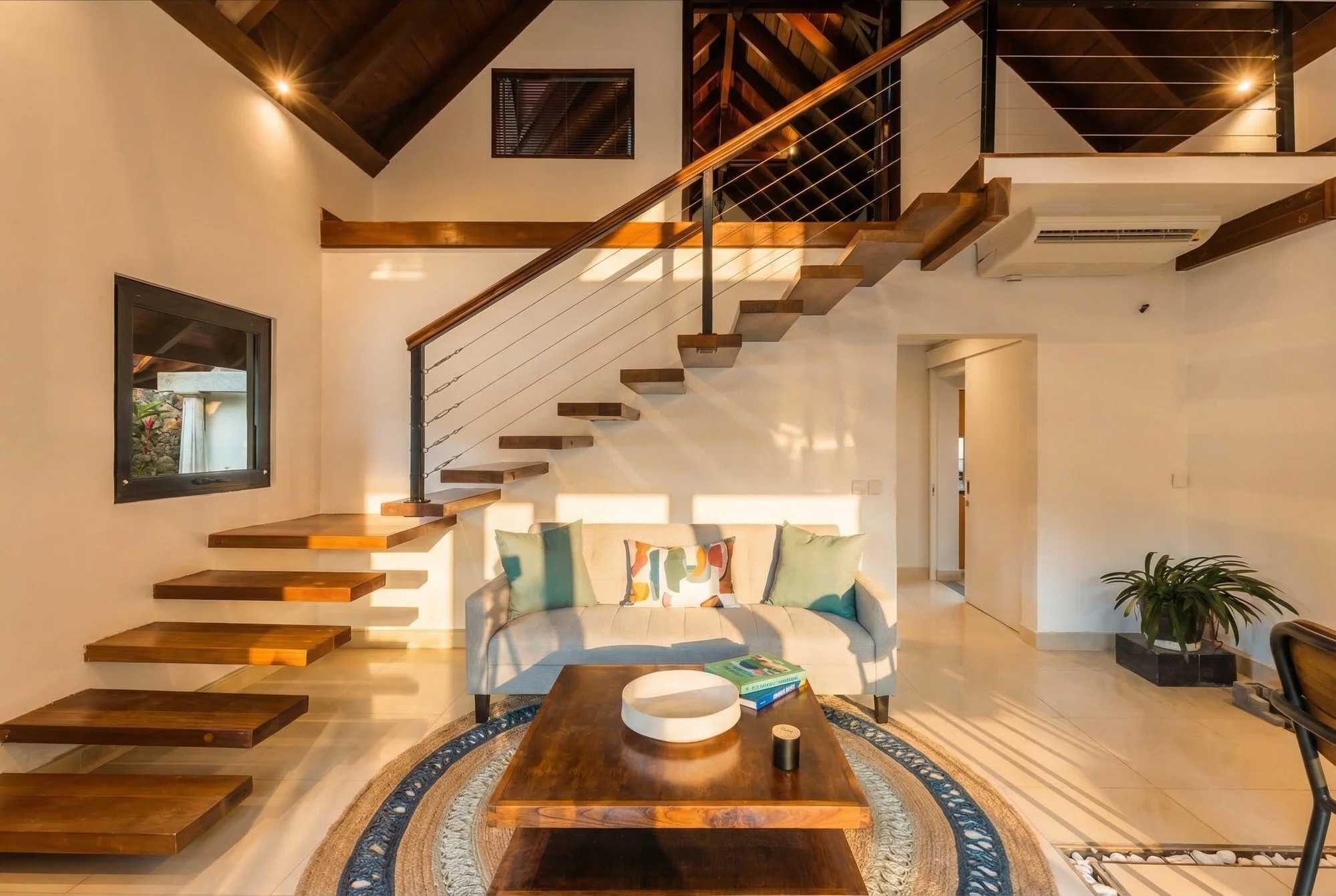 Intérieur moderne avec un escalier flottant en bois, un canapé beige avec des coussins colorés, une table en bois avec des livres, et des plantes vertes dans un espace lumineux avec murs blancs.