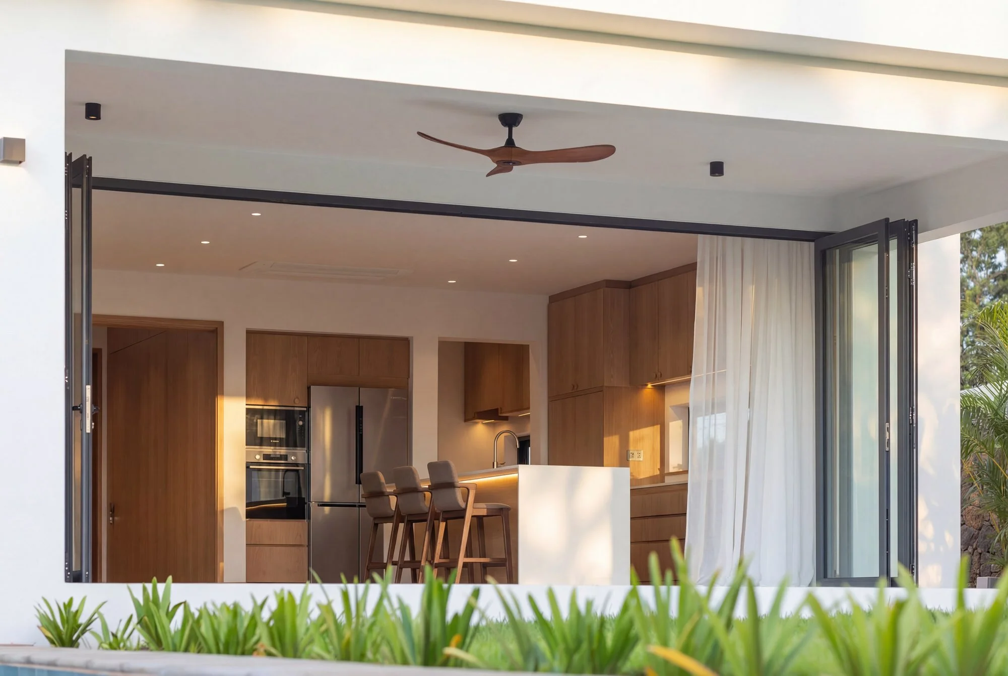 Vue intérieure d'une maison moderne avec cuisine ouverte, tables en bois, chaises, fenêtres pliantes ouvertes, plafond blanc avec ventilateur en bois.