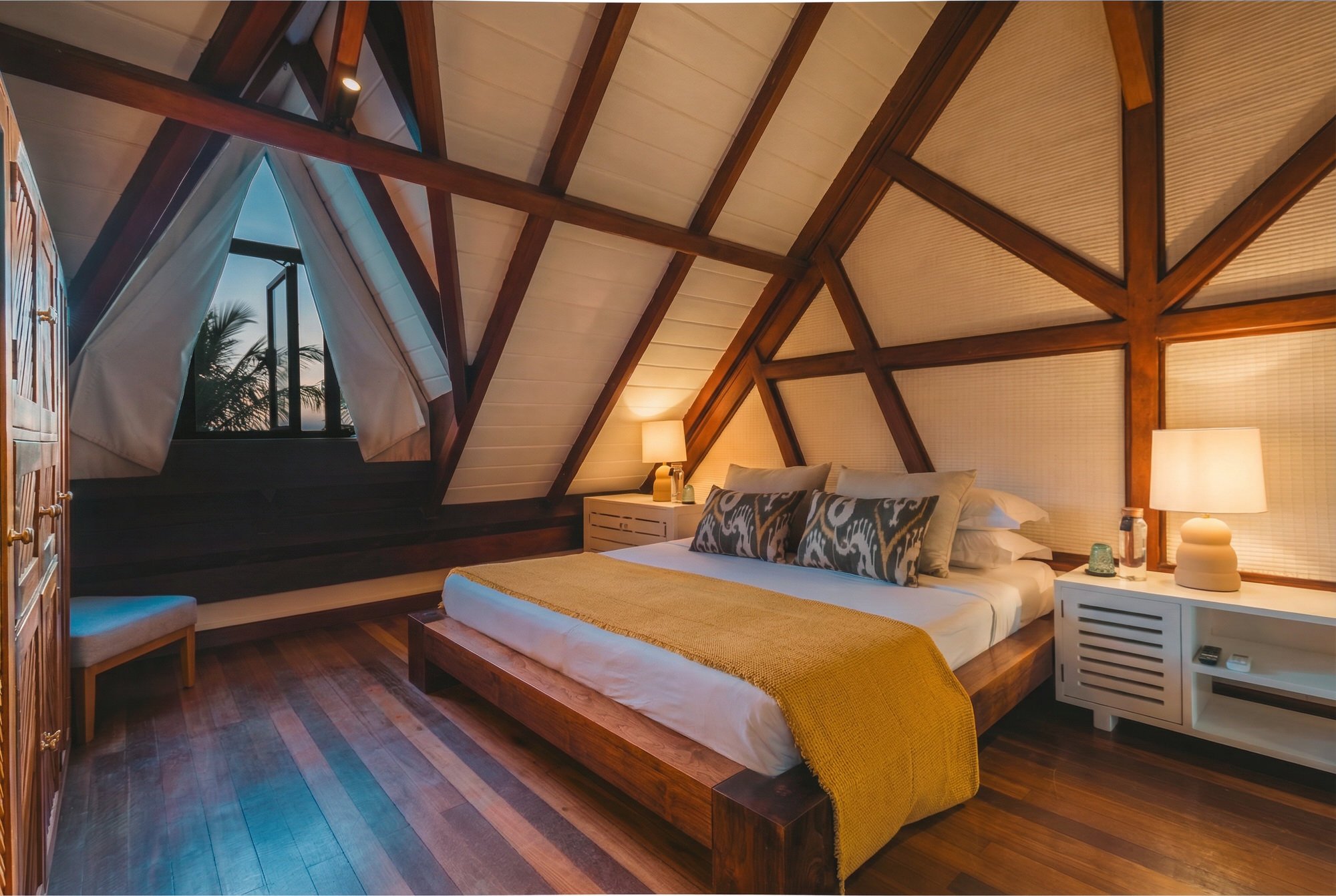 Chambre dans une cabane en bois avec un toit en pente, lit avec des oreillers à motifs, lampes de chevet, et une fenêtre laissant entrevoir un ciel clair avec des palmiers, décorée dans un style chaleureux et naturel.