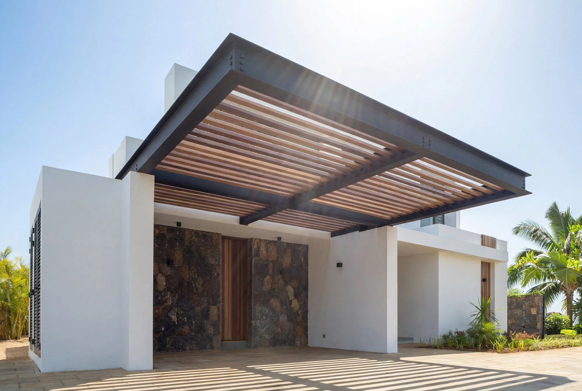 Vue d'une façade moderne d'une maison avec un auvent en bois et métal, entourée de végétation tropicale.