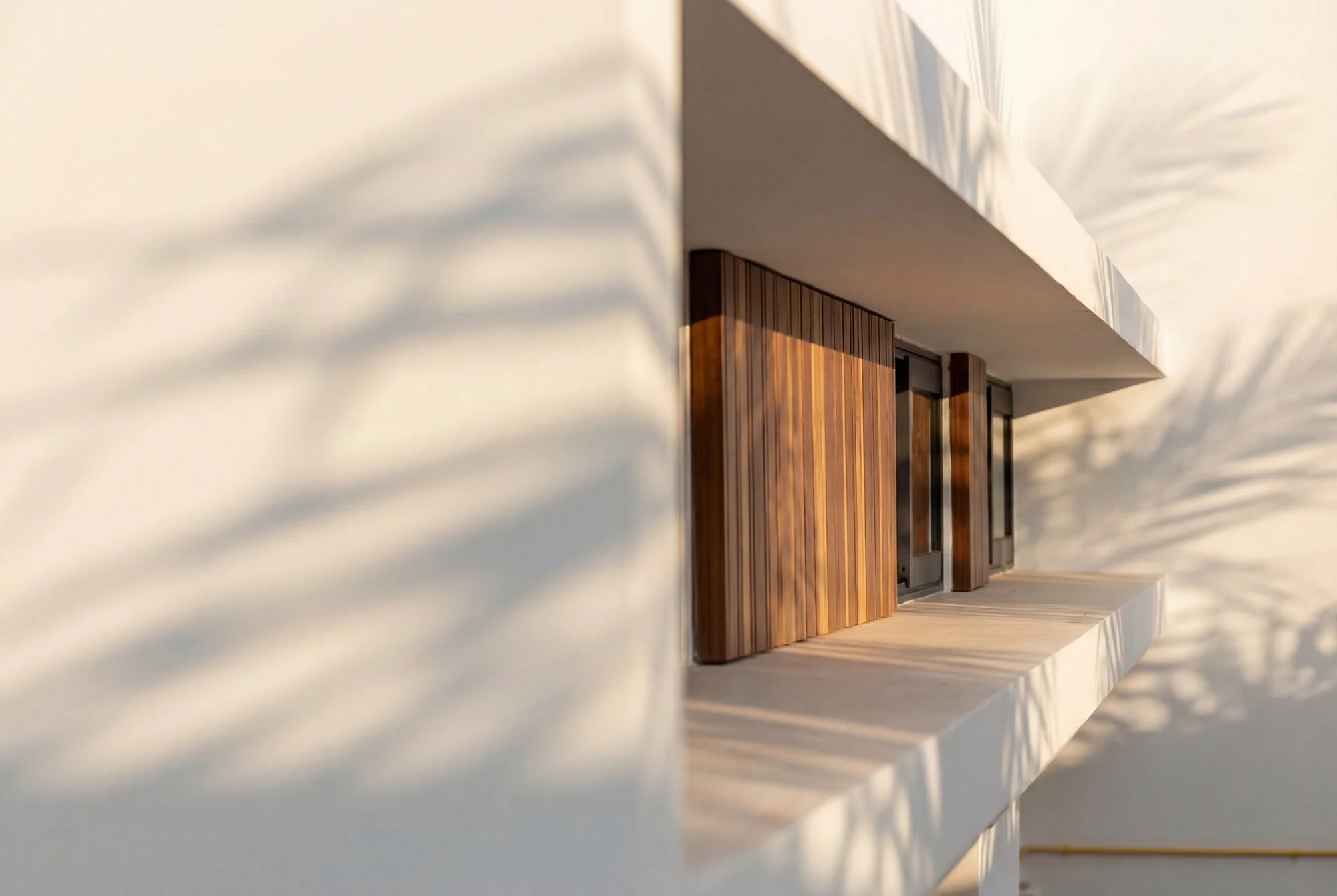 Vue latérale d'une façade moderne d'un bâtiment avec murs blancs, volet en bois et ombres portées du soleil.