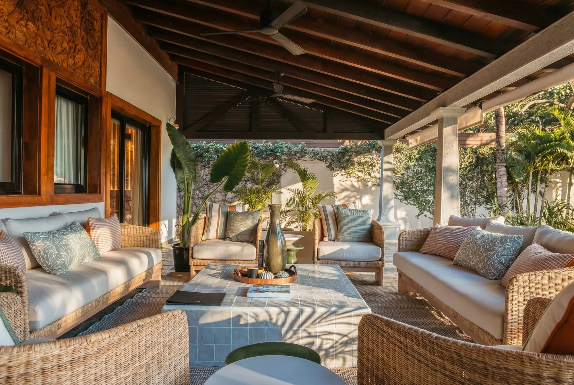 Un espace extérieur avec des canapés et des fauteuils en rotin, des coussins décoratifs, des plantes tropicales, un mur blanc, et une toiture en bois.