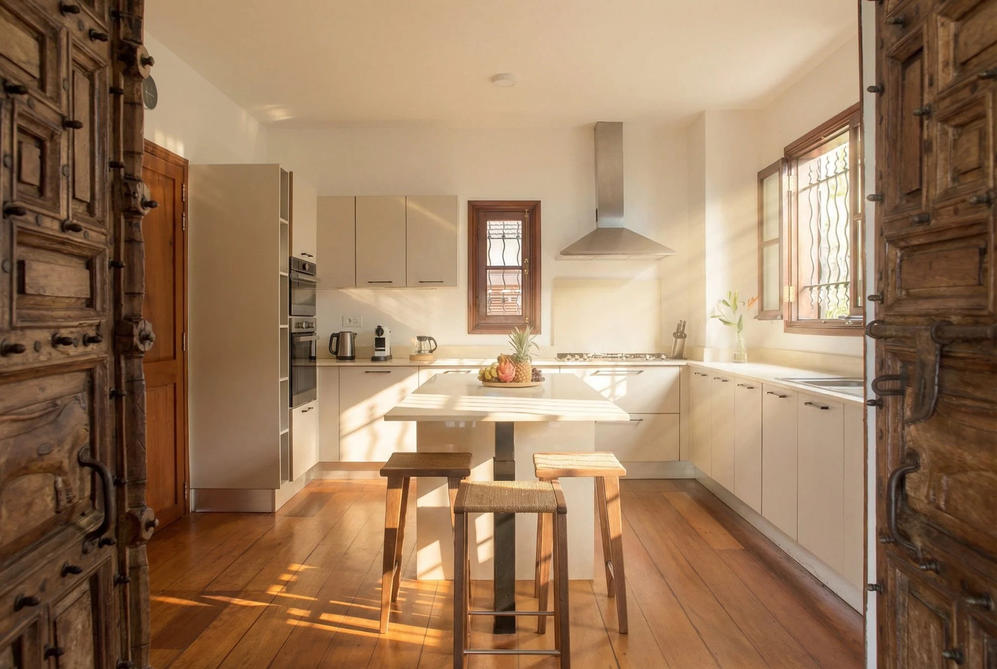 Cuisine lumineuse avec fenêtres ouvertes, mobilier moderne blanc, table centrale avec un plateau de fruits, et porte en bois ancienne à gauche.