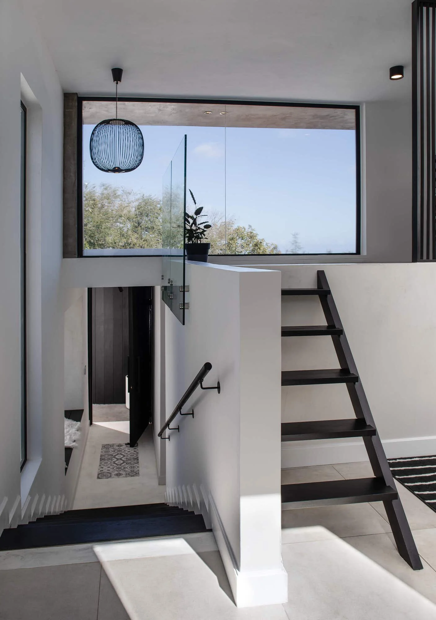 Vue intérieure d'un escalier noir avec rampe en métal, dirigé vers un espace lumineux avec une grande fenêtre laissant entrer la lumière naturelle et un pot de plante sur la balustrade