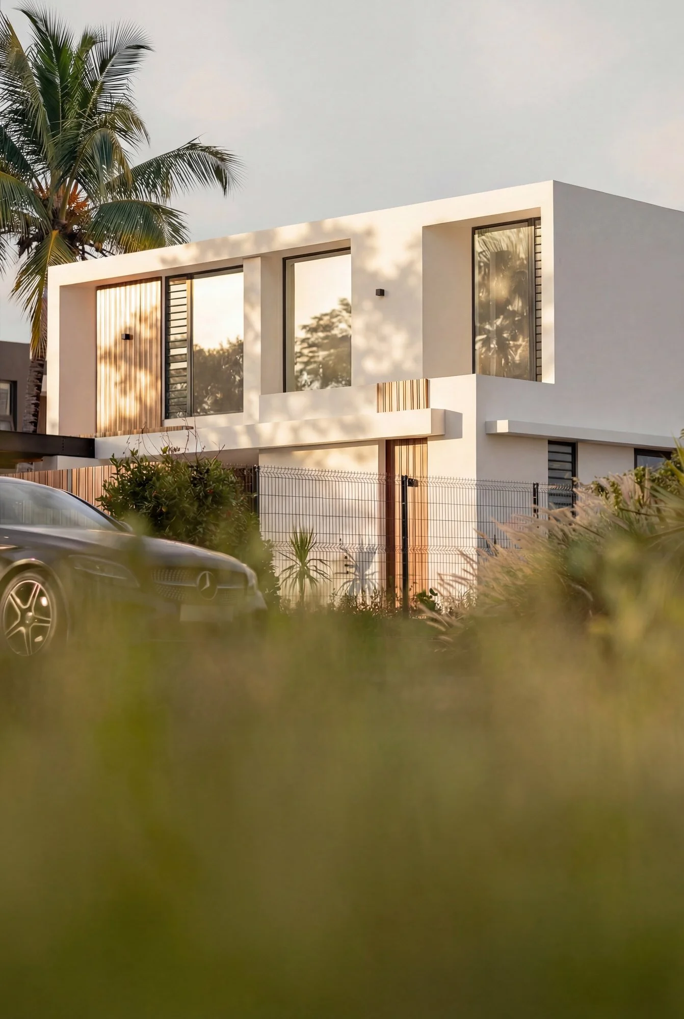 Une maison moderne blanche avec de grandes fenêtres et une porte en bois, entourée de végétation tropicale et une voiture noire garée devant, en fin d'après-midi.