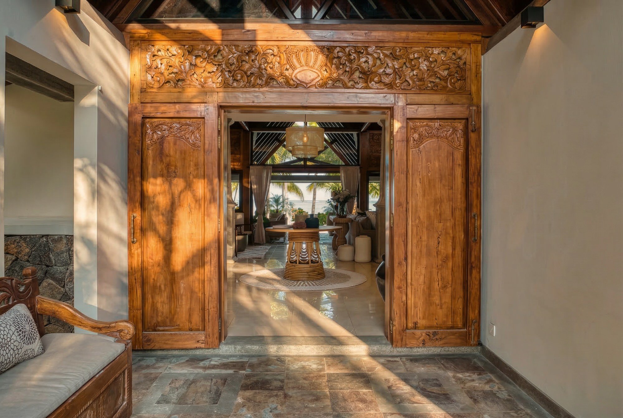 Intérieur d'une pièce avec un passage en bois sculpté, vue vers un salon avec vue sur la mer et des palmiers au fond, mobilier en bois et décoration chaleureuse.