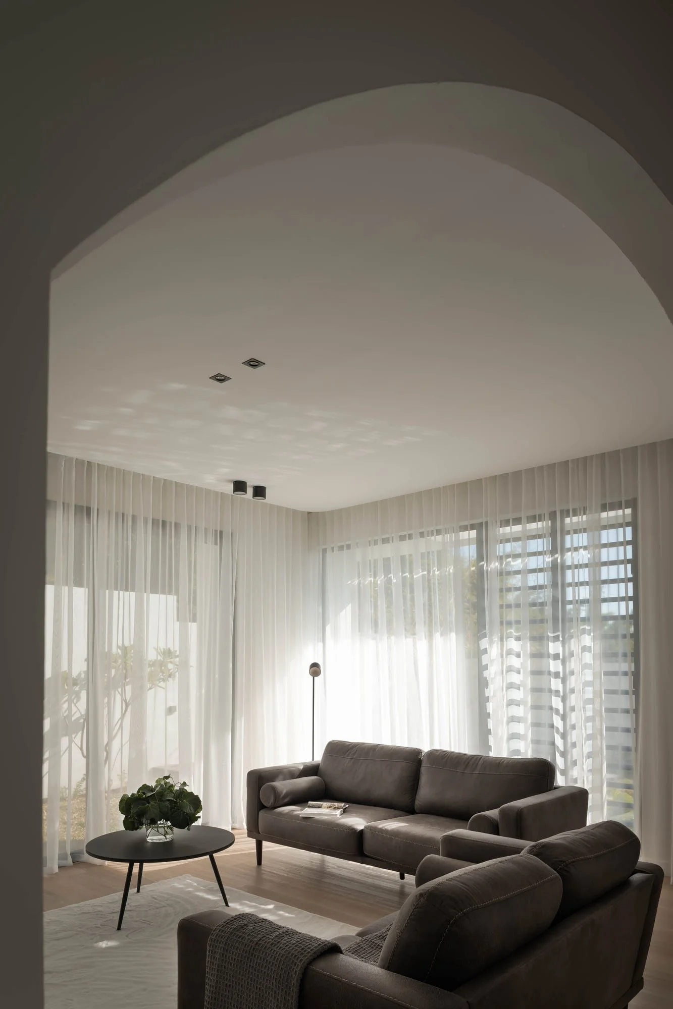 Salon moderne avec canapés gris, table basse ronde avec plante, grande fenêtre avec rideaux transparents, plafond blanc avec deux luminaires encastrés, fauteuil gris, lumière naturelle abondante.
