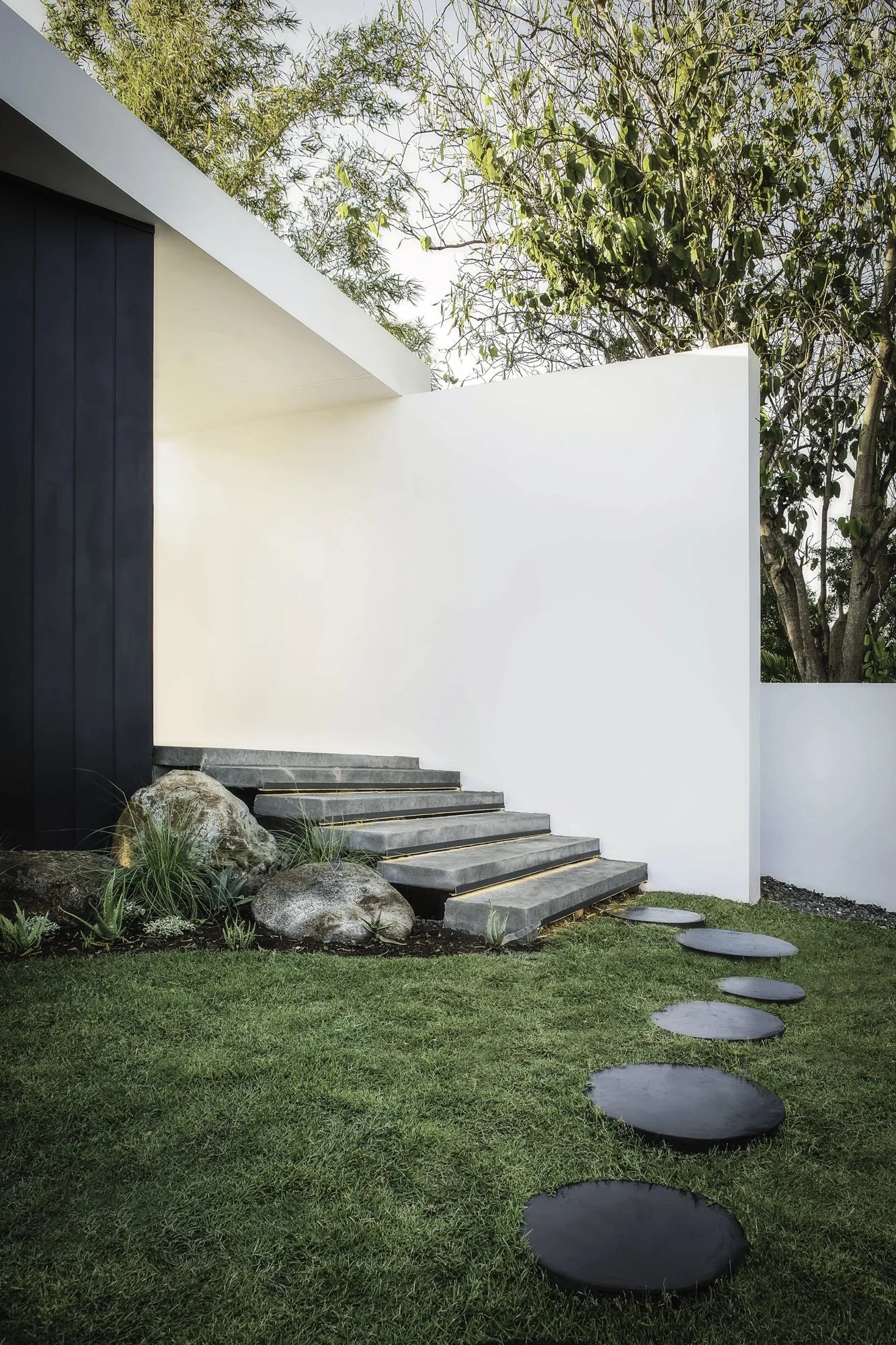 Escalier en pierre menant à une maison moderne blanche avec une façade en bois sombre, sur un jardin vert avec des pierres et des rondins en bois.