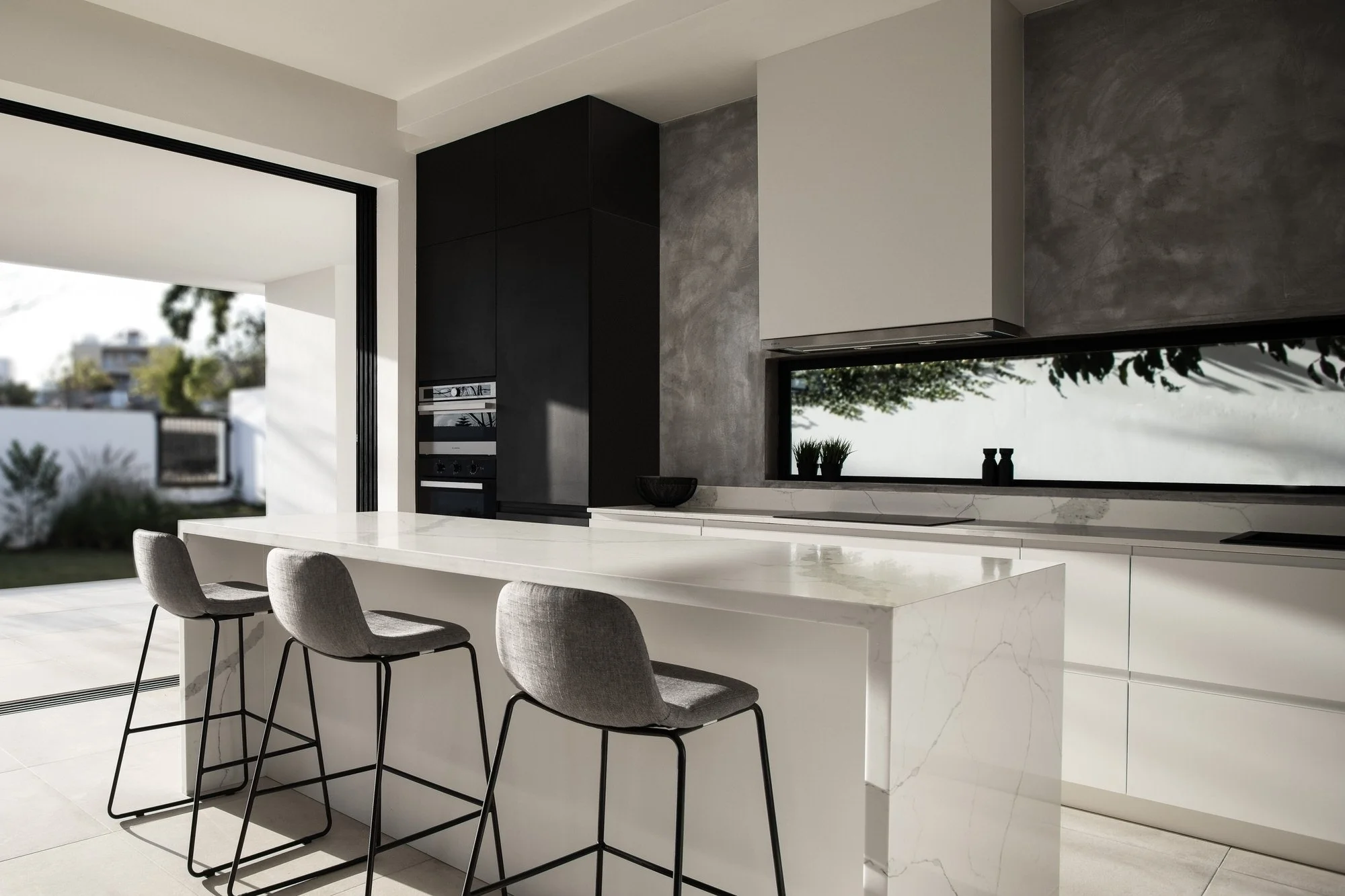 Intérieur moderne d'une cuisine avec îlot en marbre blanc, trois chaises hautes en tissu gris, grande fenêtre offrant une vue sur un jardin, murs en béton ciré et armoires noires