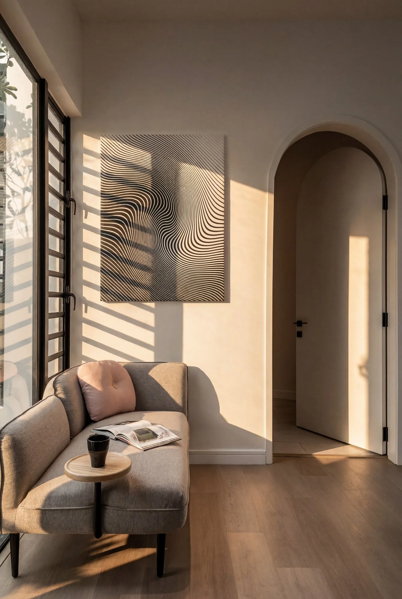 Salon moderne avec un canapé beige, un petit tableau abstrait en noir et blanc représentant un visage féminin, une fenêtre avec volets en bois laissant entrer la lumière du soleil, et un mur blanc. Un livre, un mug et une petite table en bois sont su