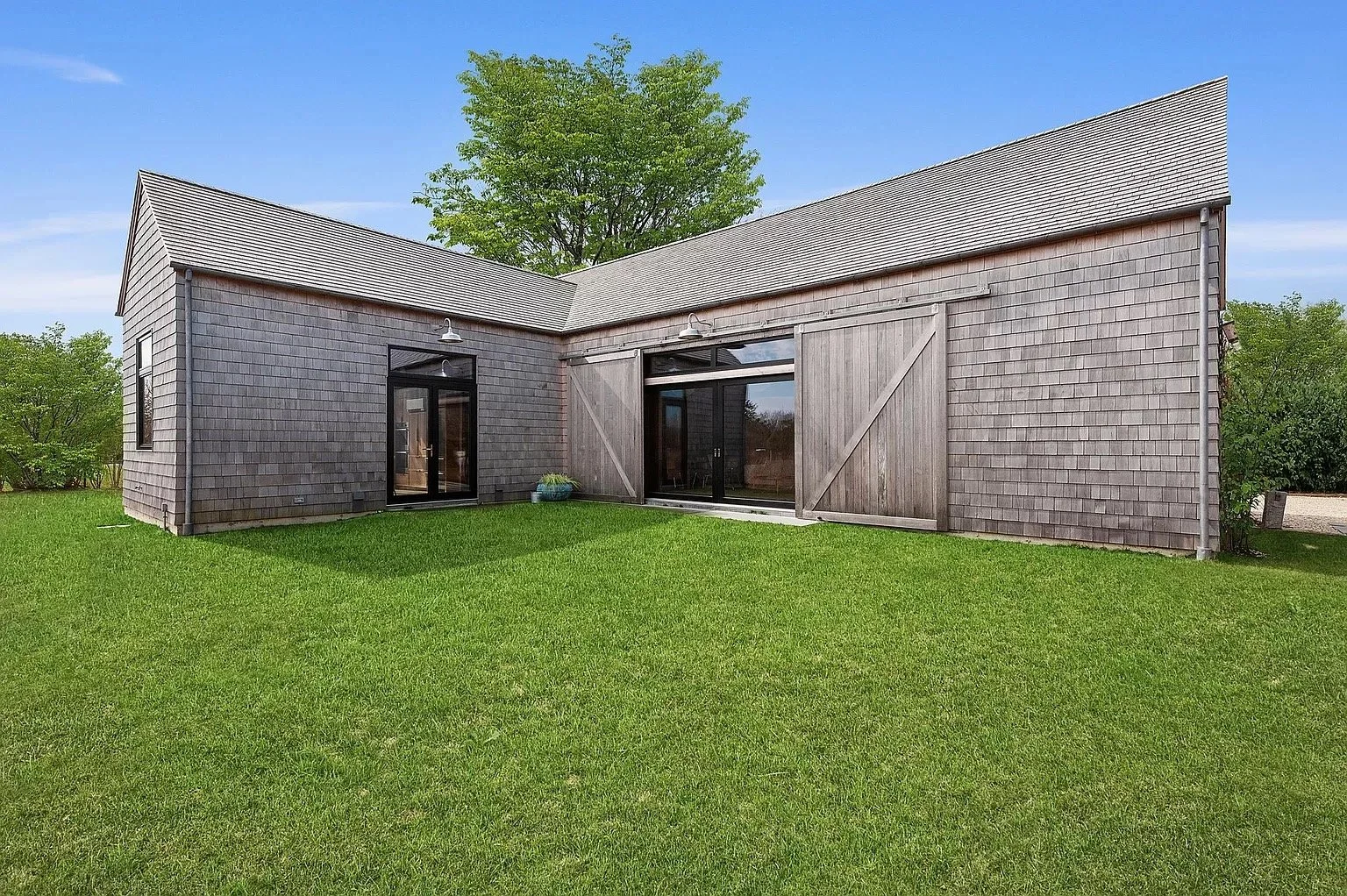 Backyard view of a house with gray wood shingles, large sliding glass doors, and a small set of steps leading to a grassy lawn, with trees and blue sky in the background.