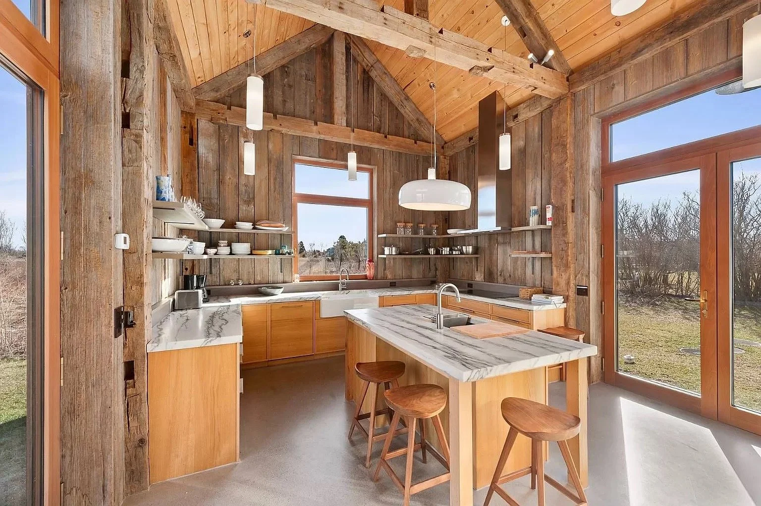 Modern rustic kitchen with wooden walls, ceiling, and cabinets, large windows, a marble island with stools, and open shelves.
