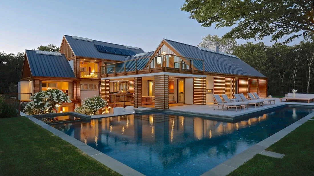 Modern house with wooden siding and a metal roof, featuring solar panels, large windows, and a balcony, located near a swimming pool with poolside lounge chairs. Lush trees surround the property at dusk.