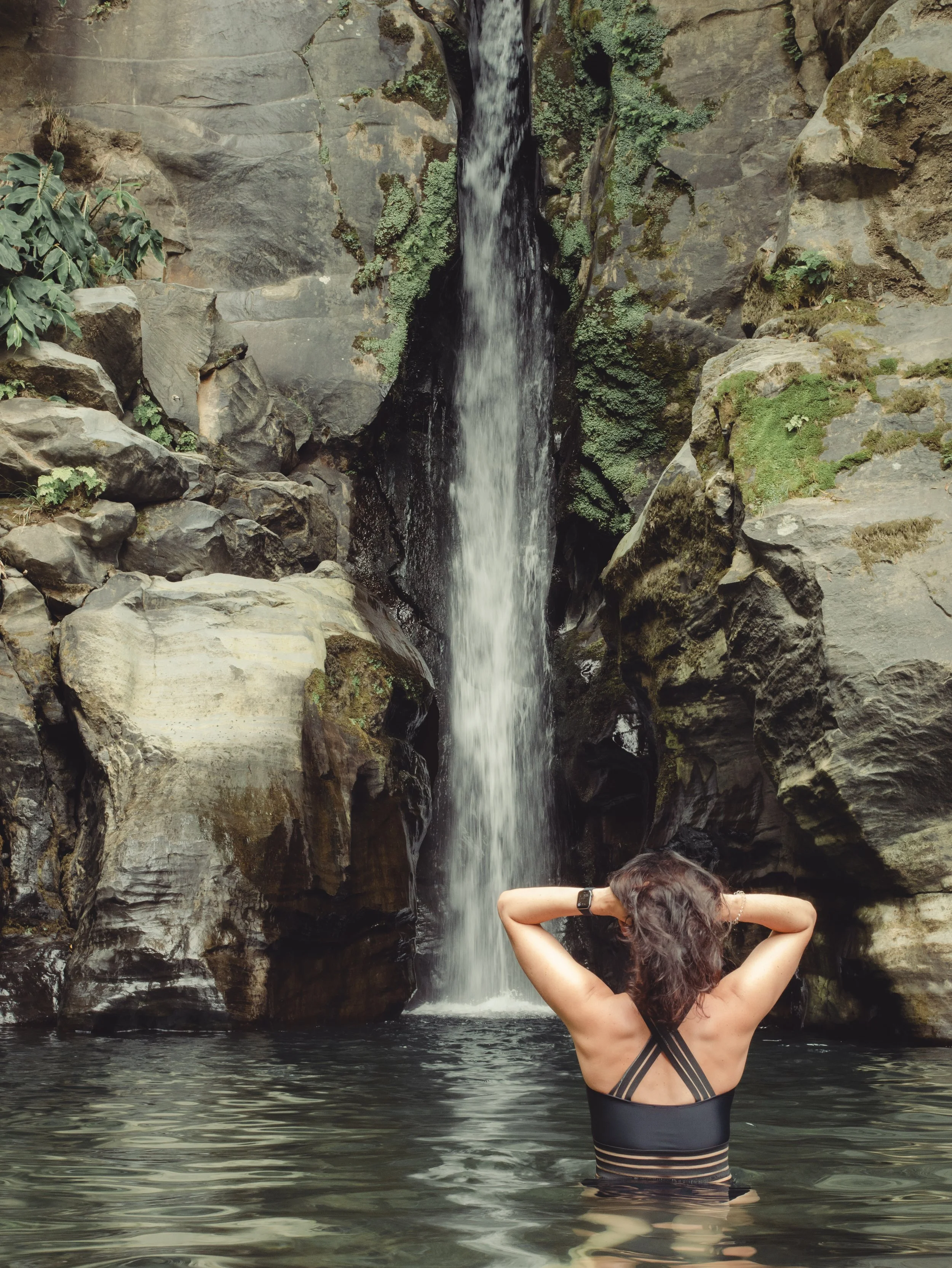 Woman in a black swimsuit relaxing in the clear pool beneath a natural waterfall in São Miguel, Azores. Female portrait captured during a solo photo session.