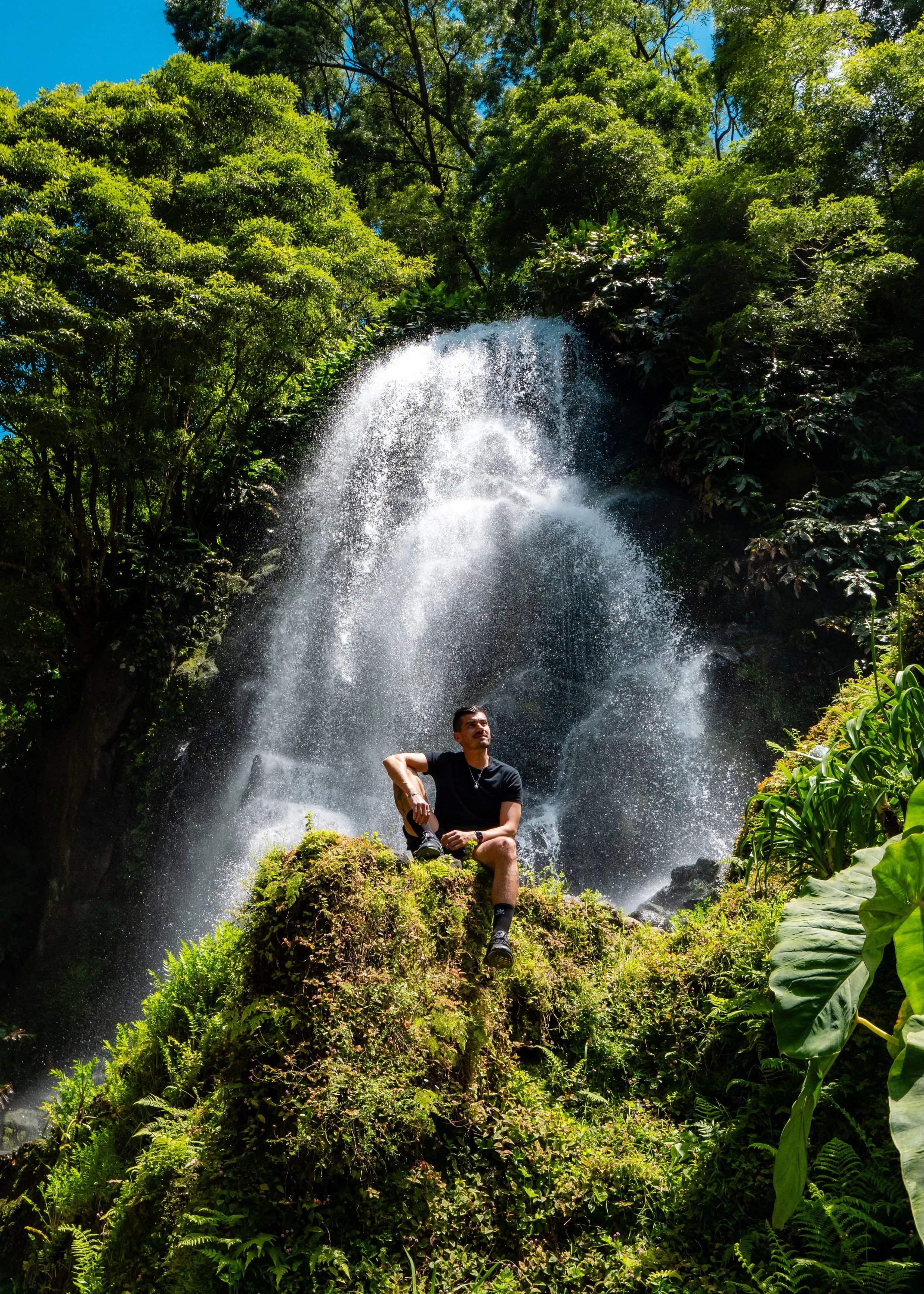 Portrait of a man sitting on bright green moss in front of a large, lush waterfall in São Miguel, Azores. Focus is on soft natural light and the water's movement