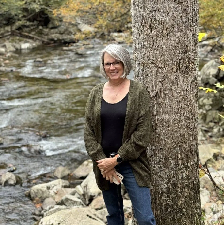 A woman with gray hair and glasses standing outdoors beside a tree next to a rocky creek, smiling and holding a phone.
