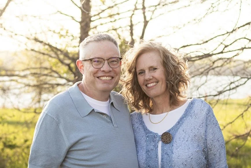 A smiling man and woman standing outdoors in front of a tree during sunset.