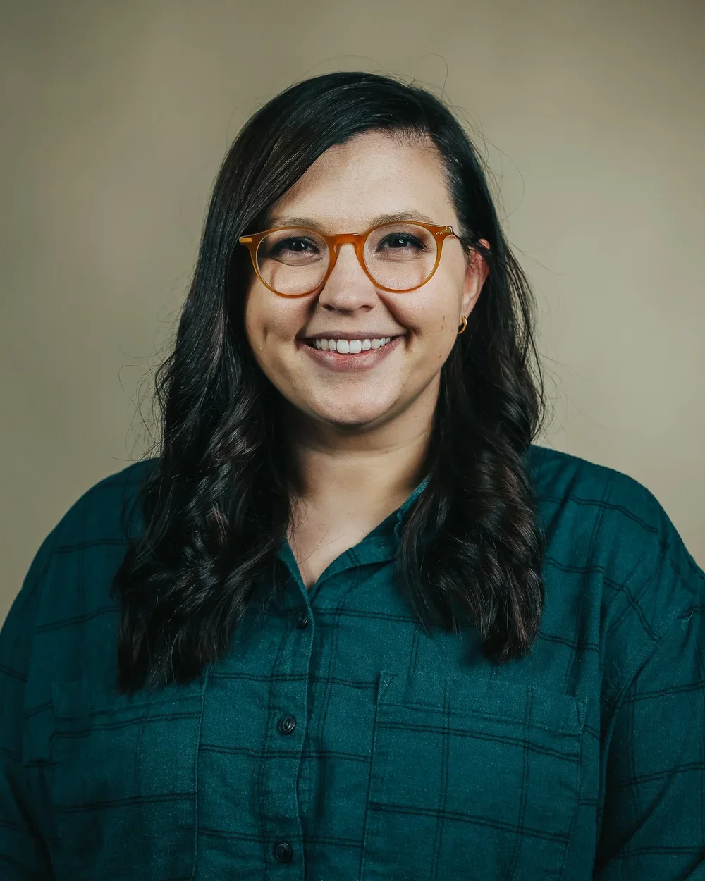 A woman with long dark hair, wearing glasses and a teal flannel shirt, smiling at the camera against a neutral background.