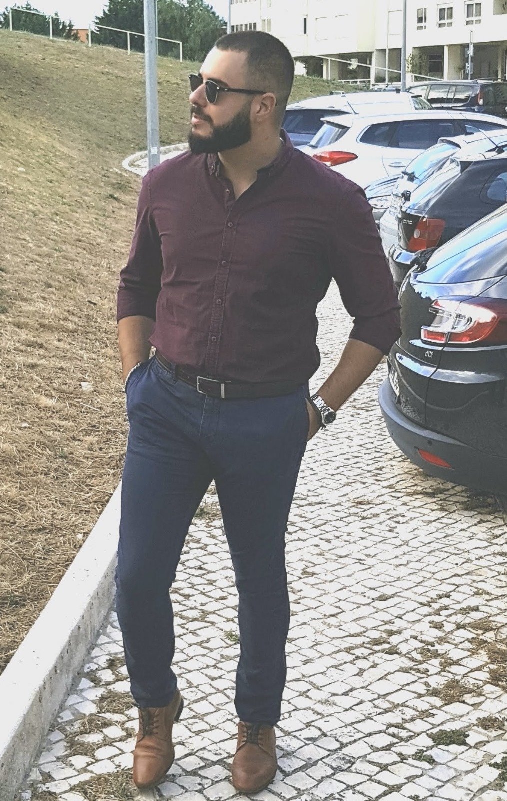 Man with sunglasses and beard standing on cobblestone path near parked cars.