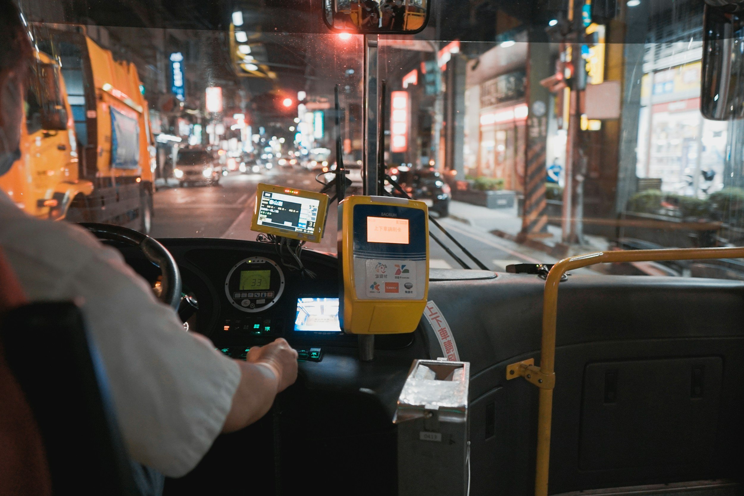 Interior of a bus at night, showing a driver's seat, steering wheel, digital display screens, fare box, and city street lights visible through the windshield.