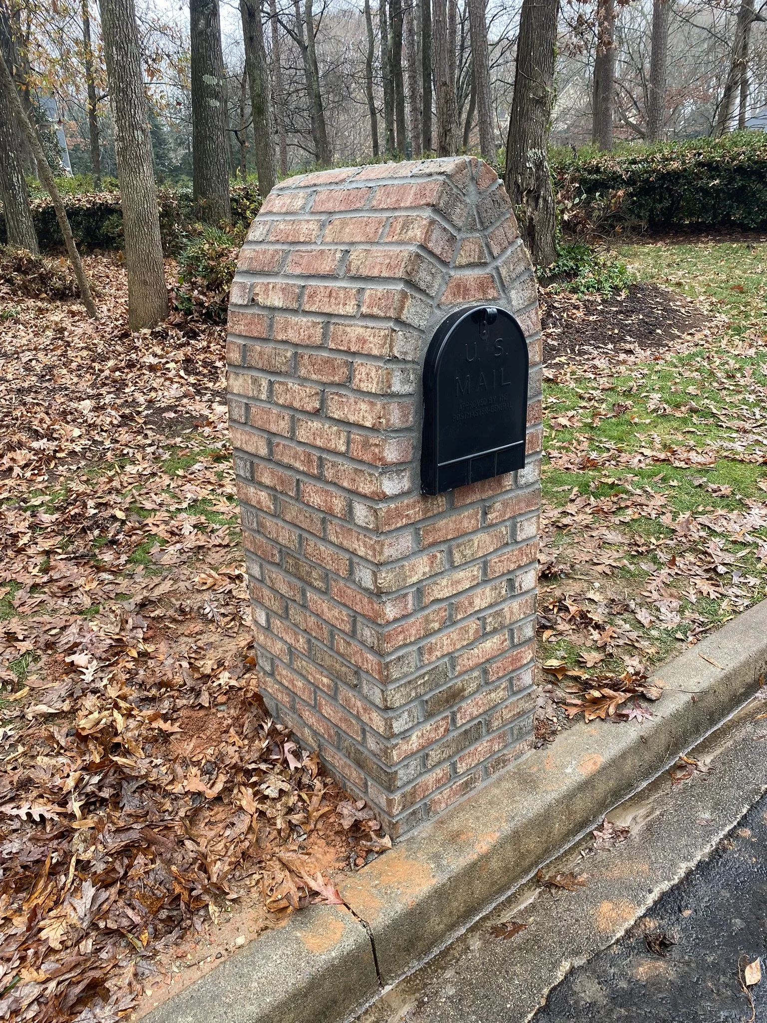 A brick mailbox with a black mailbox attached, set on a concrete sidewalk, surrounded by fallen autumn leaves and trees in the background.