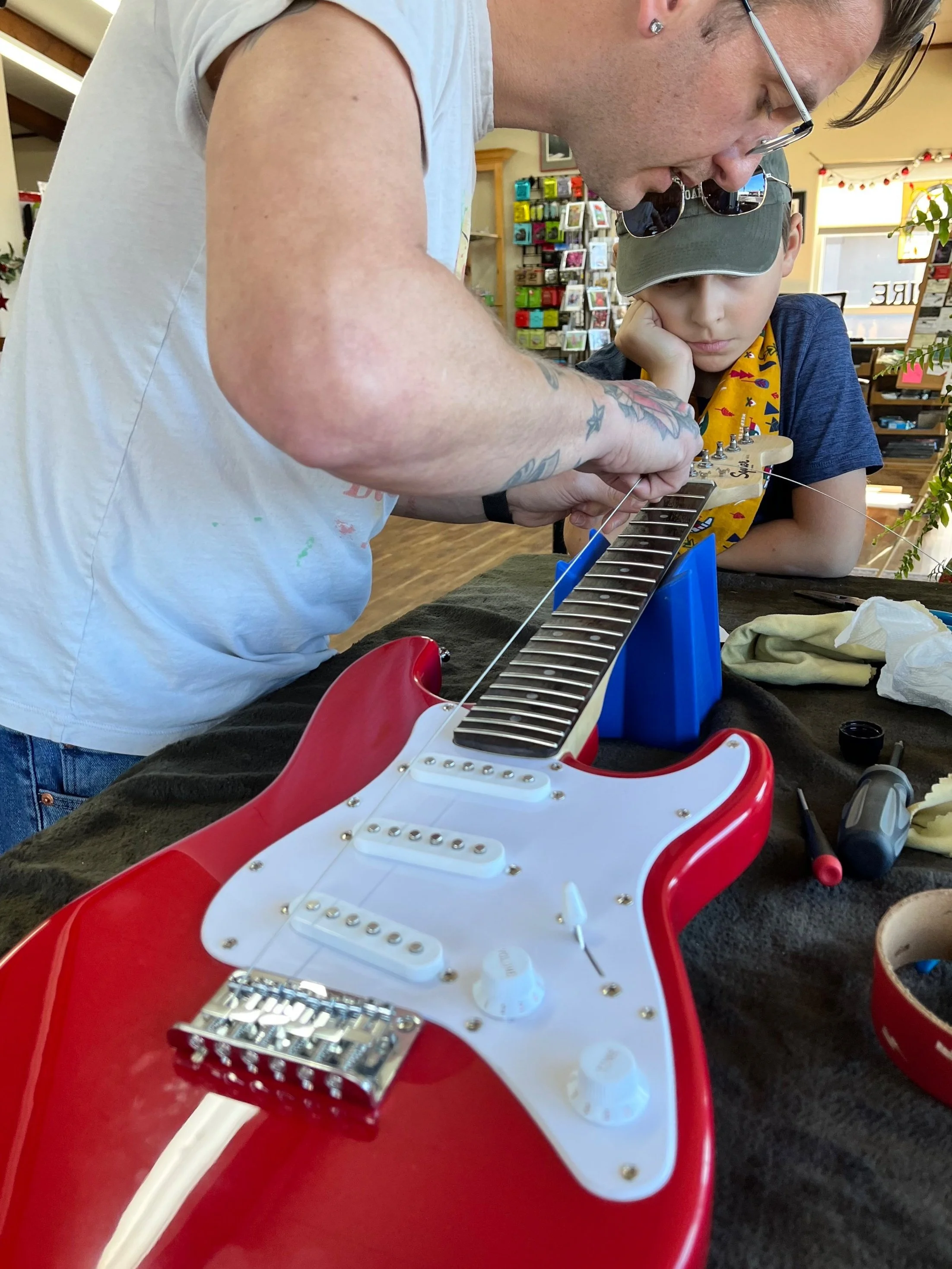 Person adjusting strings on a red electric guitar with a child observing.