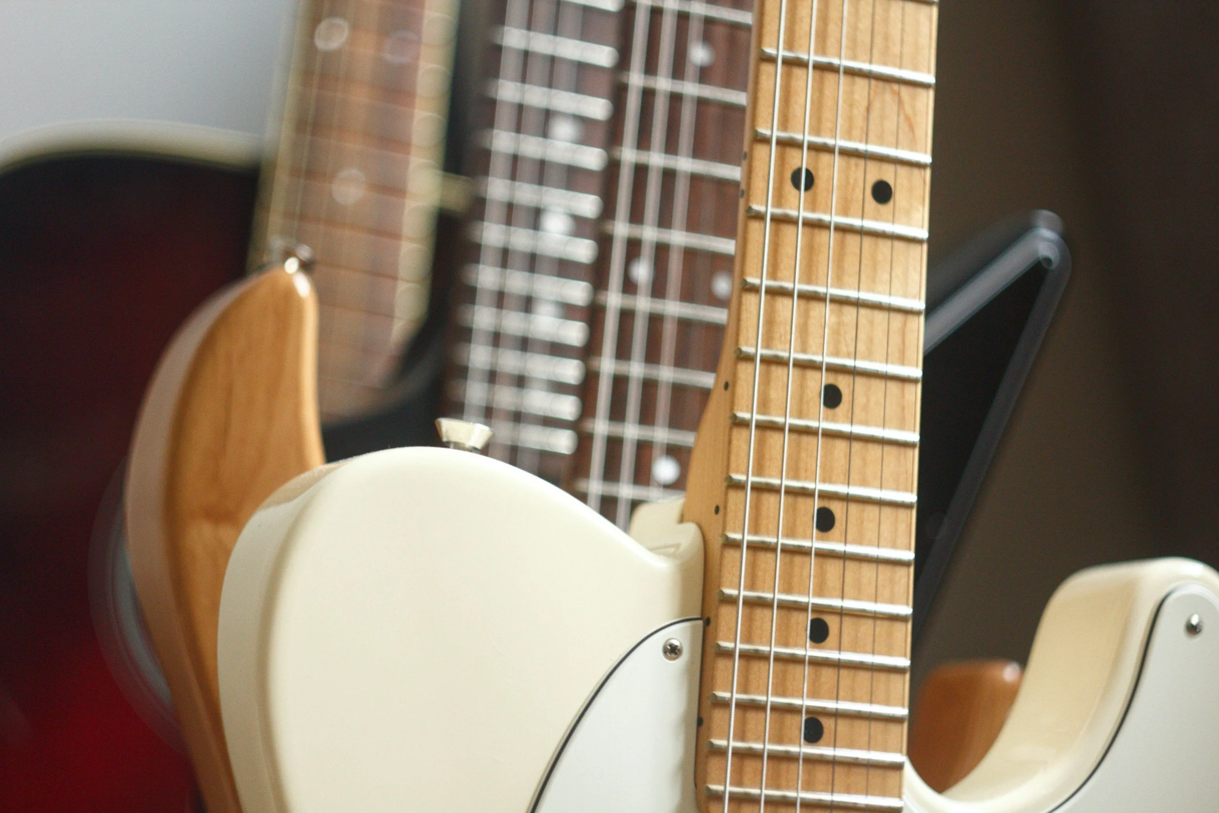 Close-up of a cream-colored electric guitar with a wooden neck, multiple guitar necks in the background, some with darker wood and metal strings.