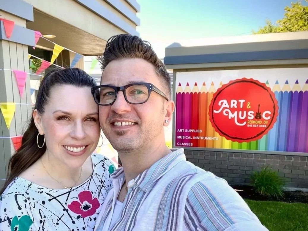 A smiling couple taking a selfie in front of an art and music store advertising art supplies, musical instruments, and classes, with colorful bunting in the background.