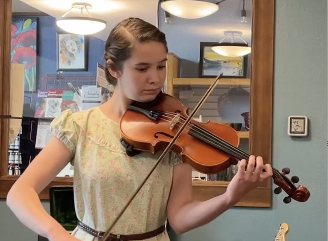 Young woman playing a violin indoors, standing in front of art and books.