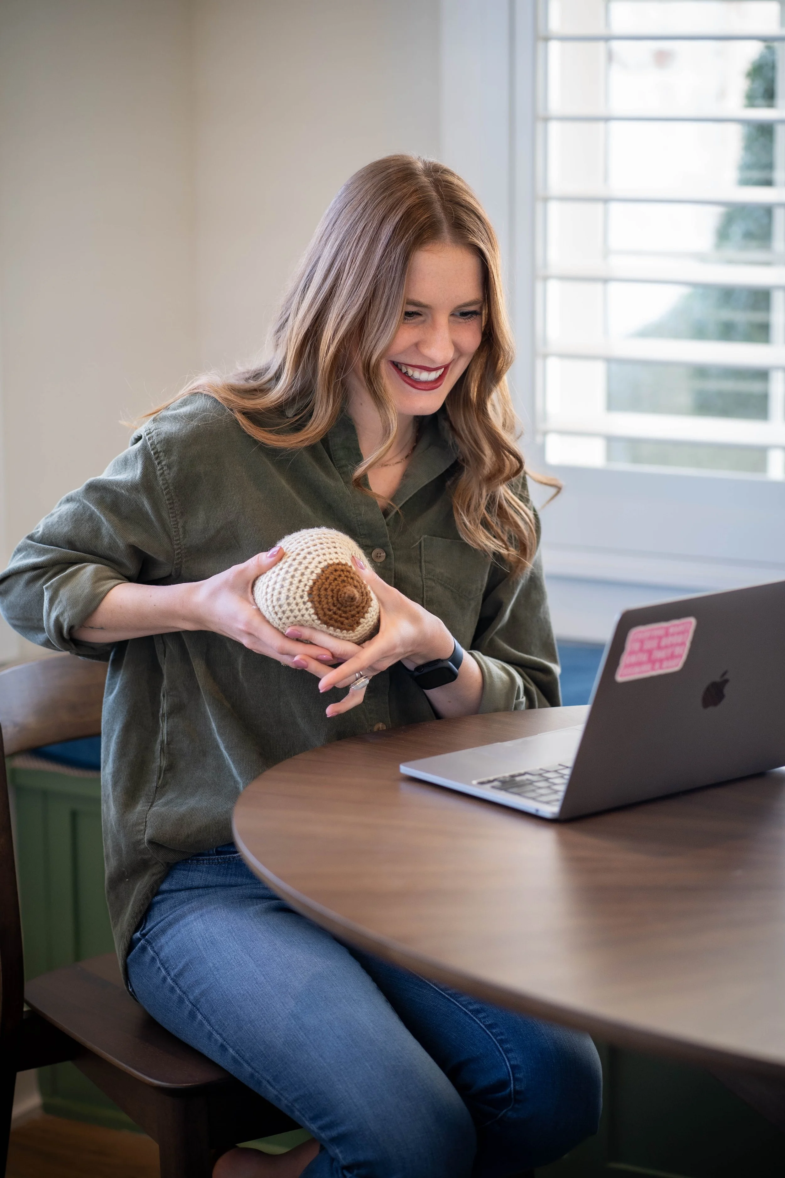 A woman with long light brown hair, wearing a green jacket and blue jeans, sitting at a round table using a silver MacBook laptop, holding a crochet item, smiling inside a brightly lit room with a large window.