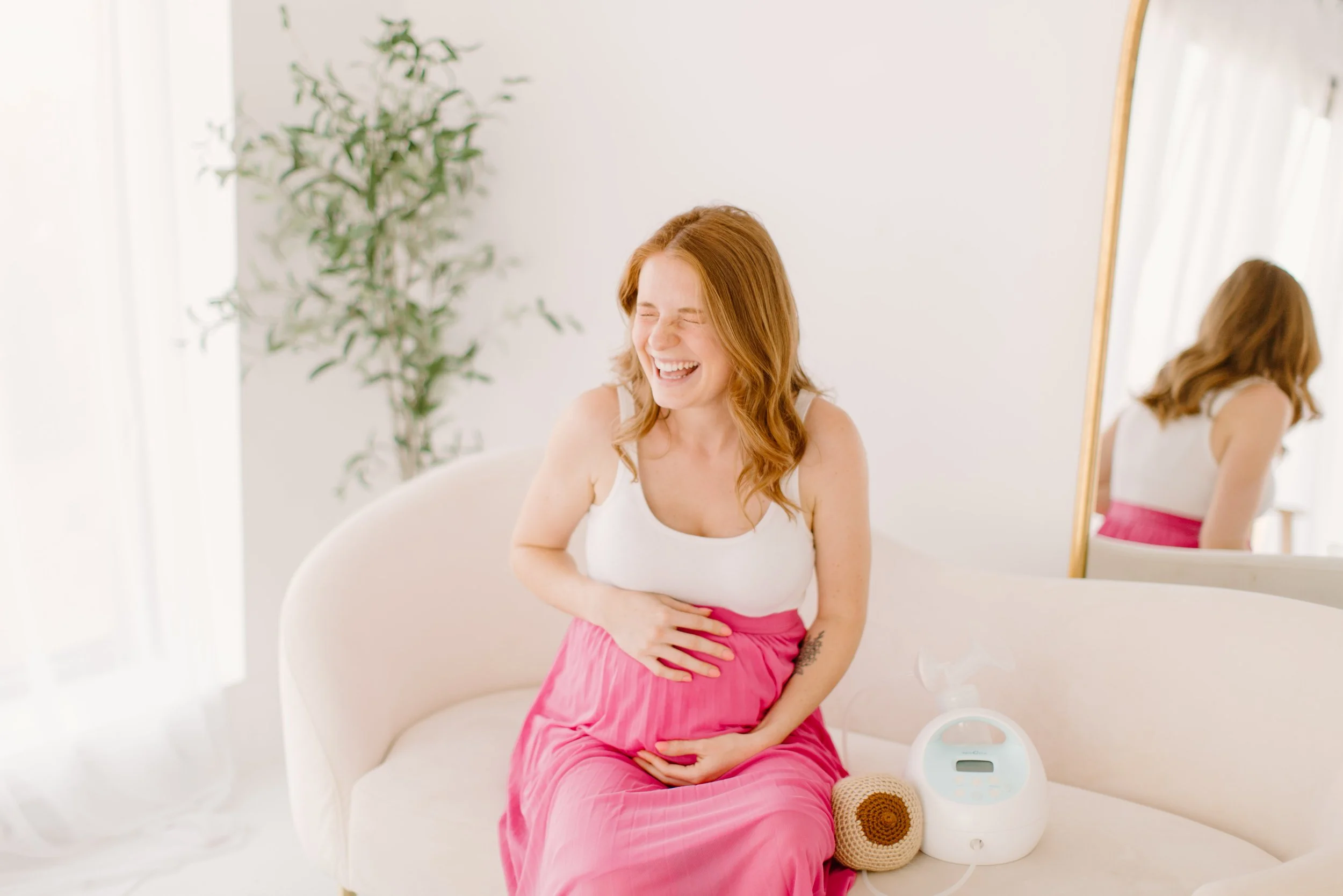 A woman with red hair laughing while sitting on a white sofa, holding her stomach with both hands. She is wearing a white tank top and a pink skirt. There is a mirror behind her, reflecting her back. A green plant is in the background, and a white device is on the side of the sofa.