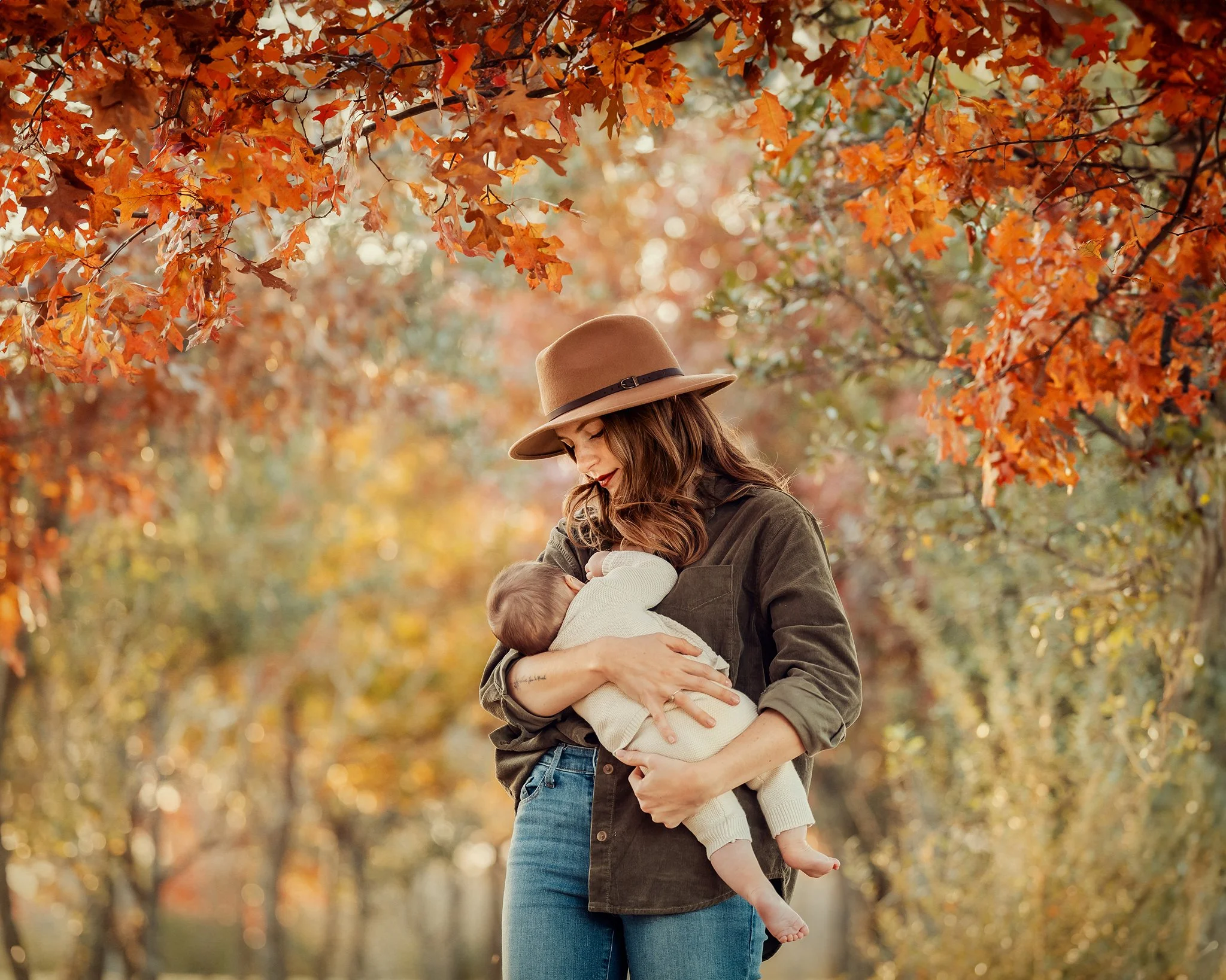 A woman with long brown hair, wearing a tan hat and a green jacket, holding a young child with blonde hair, dressed in a white sweater, in an outdoor setting with colorful autumn leaves overhead.