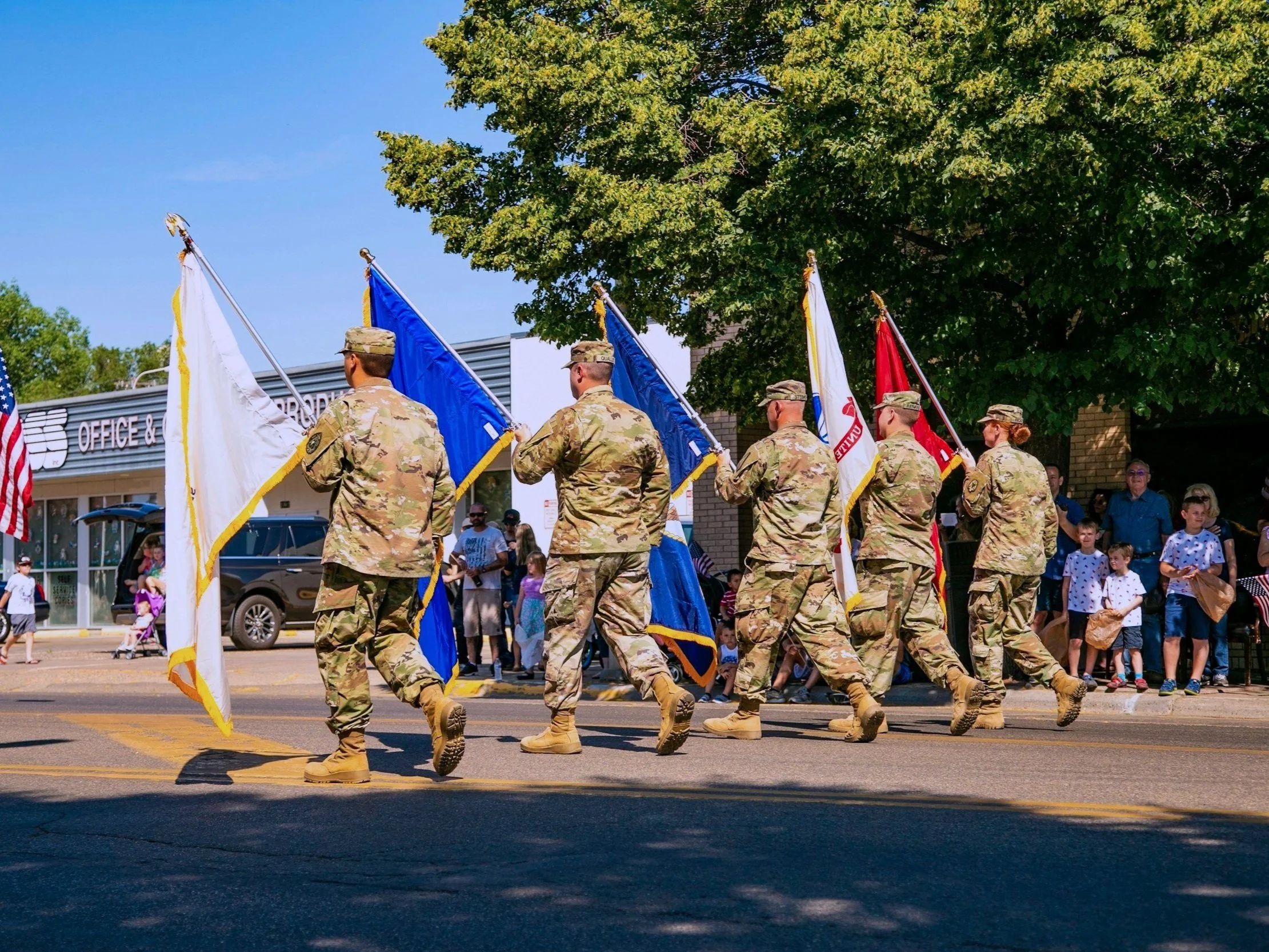 Members of the US military march in color guard at a parade on a street in summer.
