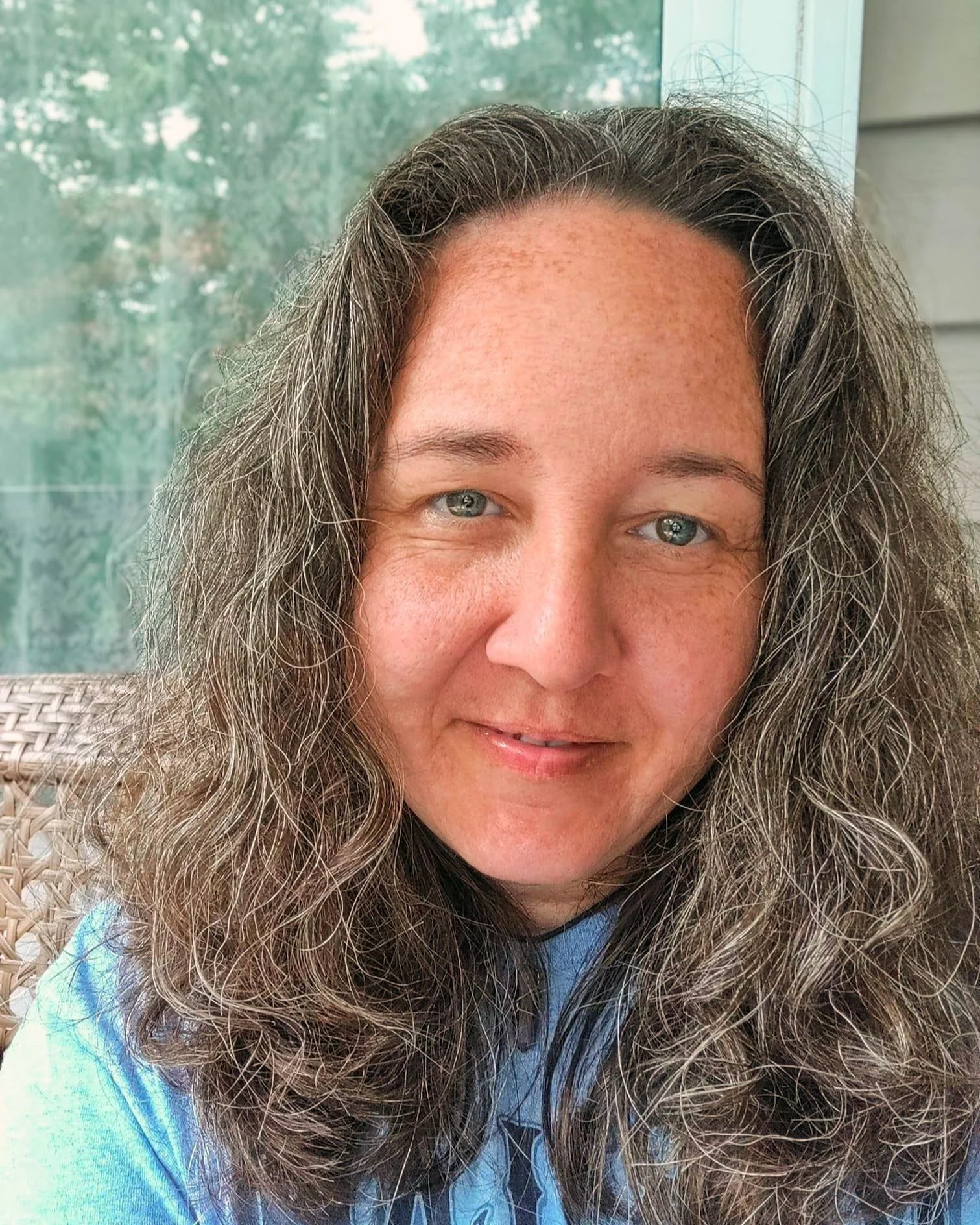 Melissa Cantara looks into the camera. She has brown hair with gray strands, hazel eyes, and a kind face with light skin and freckles. She is wearing a blue shirt with navy lettering and sitting in a wicker chair.